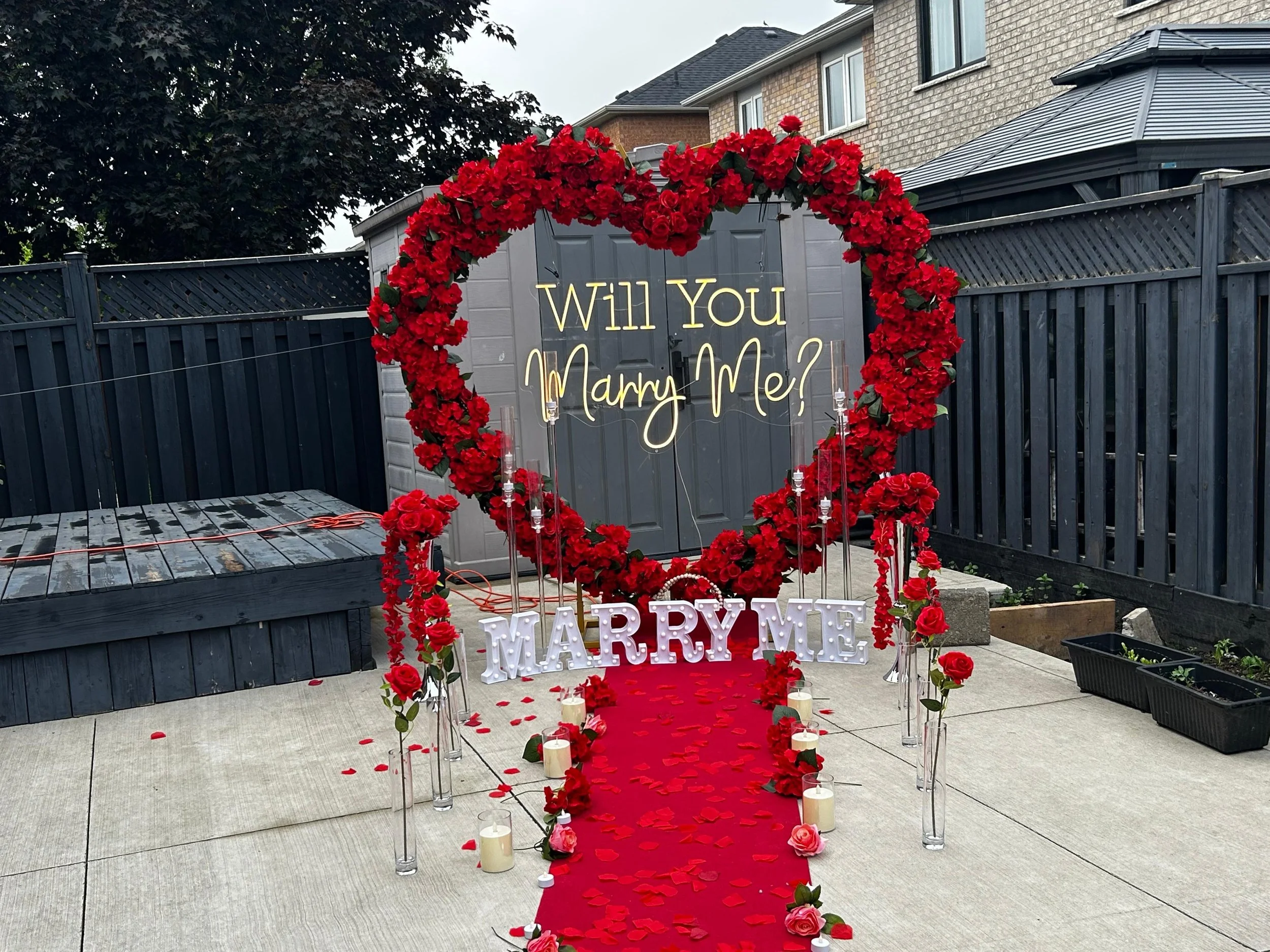 A romantic wedding proposal setup outdoors featuring a heart-shaped arch decorated with red roses, a red rose petal-covered aisle, candles, and a sign asking 'Will You Marry Me?' with additional 'MARRY ME' letters at the end of the aisle.