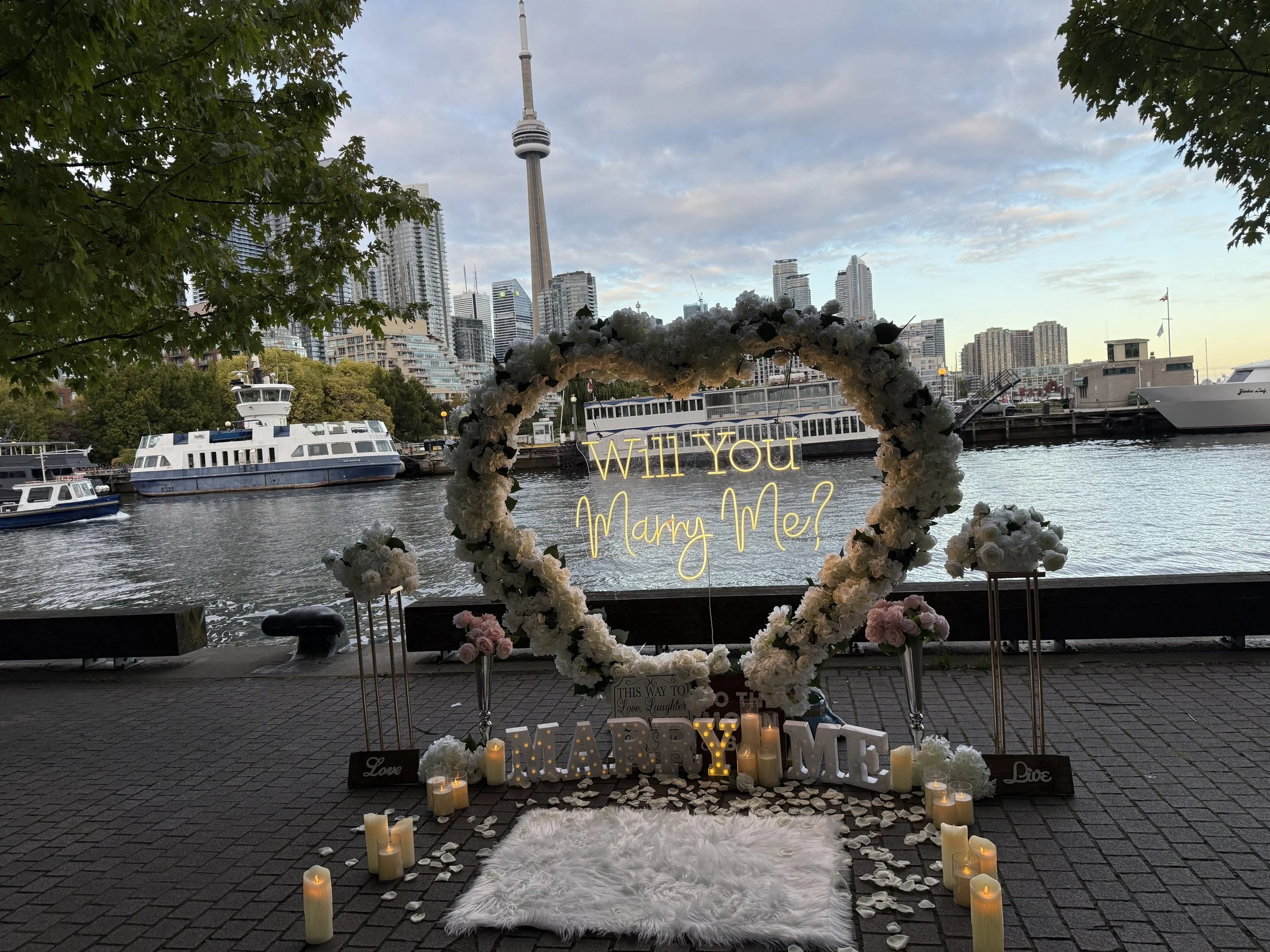 Proposal setup near a river with a city skyline background. The arrangement features a heart-shaped floral arch, candles on the ground, and illuminated signs that read "Will You Marry Me?" with additional decorative elements.