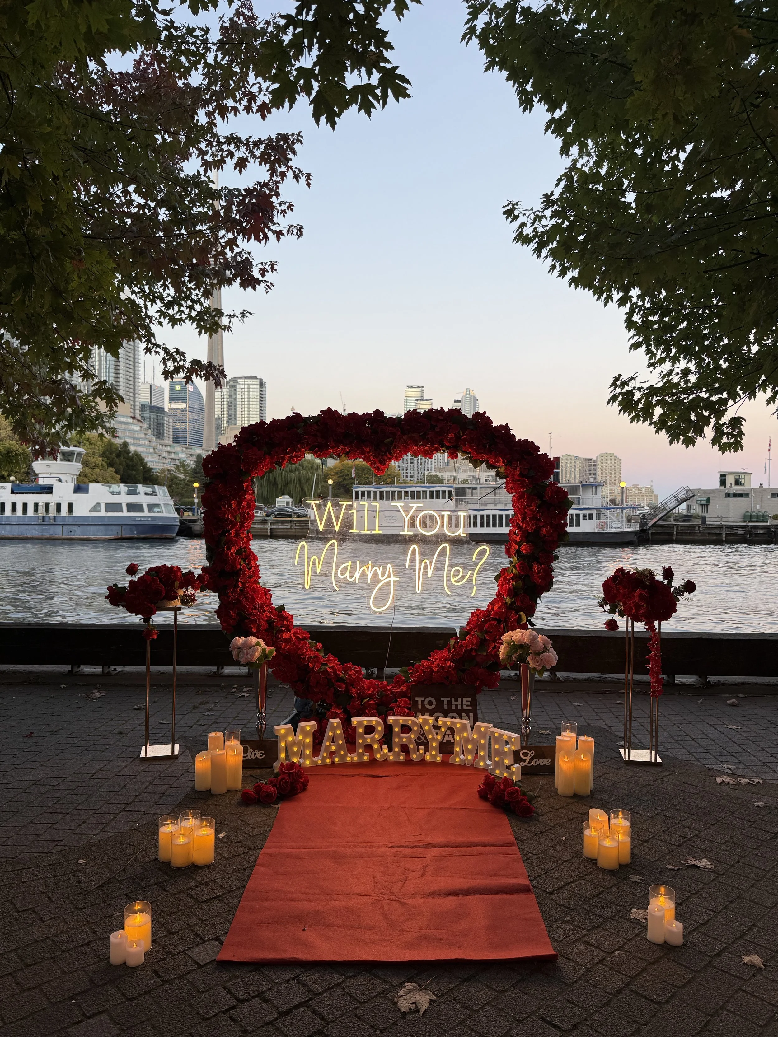A romantic proposal setup by a waterfront with a heart-shaped floral arch, a neon sign reading 'Will You Marry Me?', candles, flowers, and a red carpet leading up to the message, with boats and city skyline in the background at sunset.