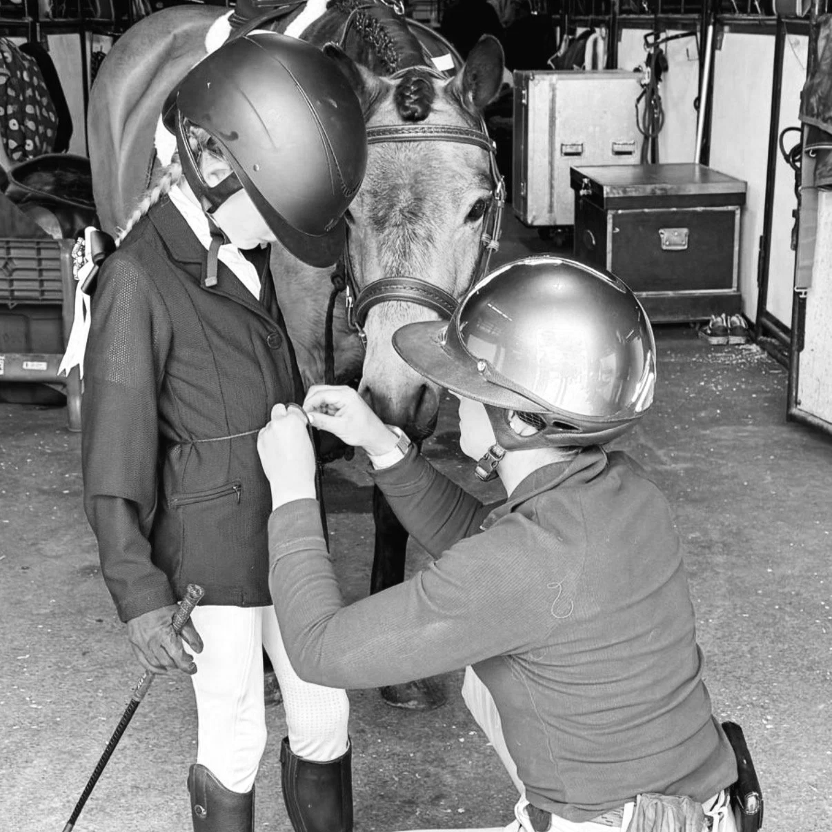 A person in riding gear is being fitted with a medal or ribbon by another person, both wearing helmets, while a horse looks on in a stable or tack room.