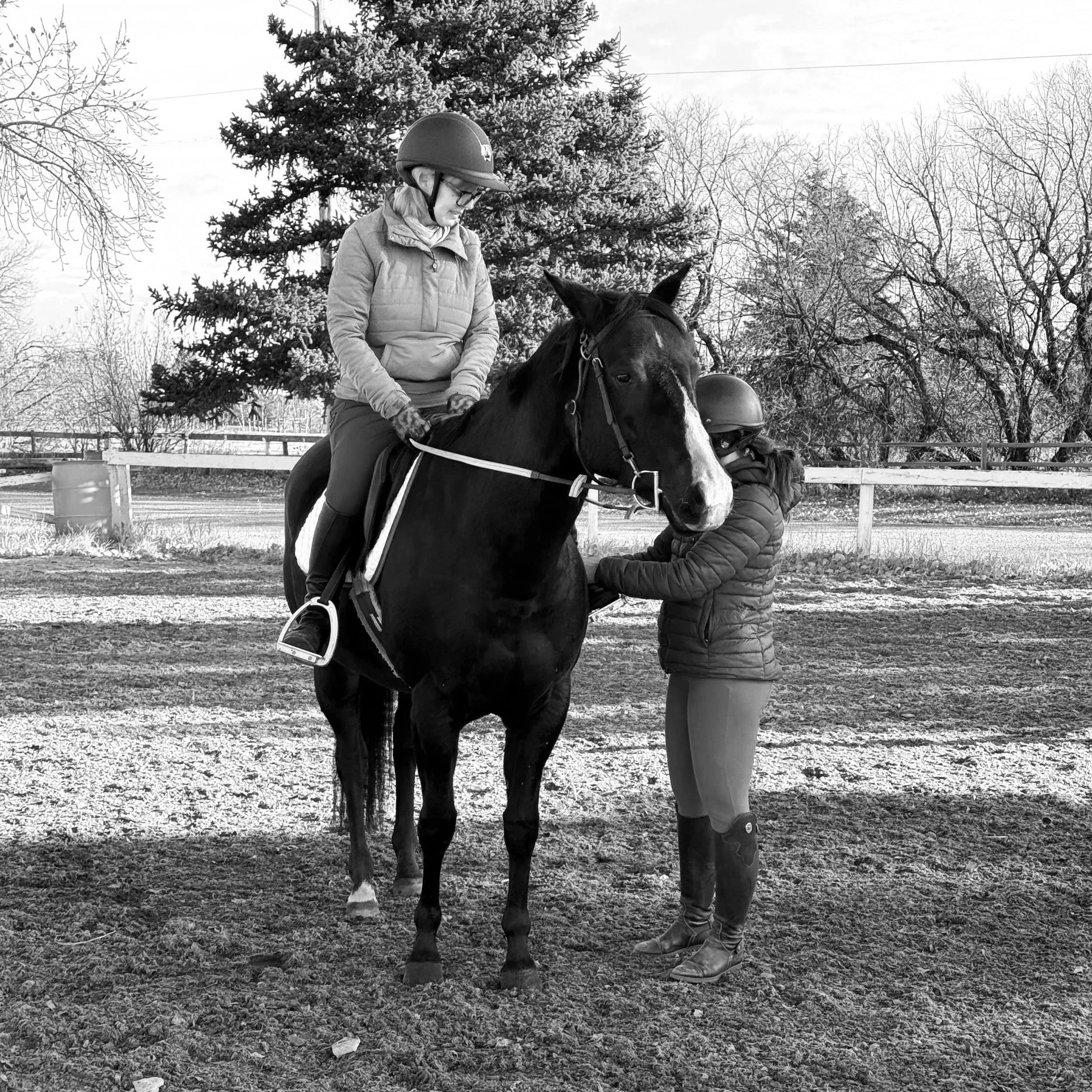 A woman wearing a helmet and glasses riding a horse, with a child standing next to the horse holding its reins, outdoors with trees in the background.