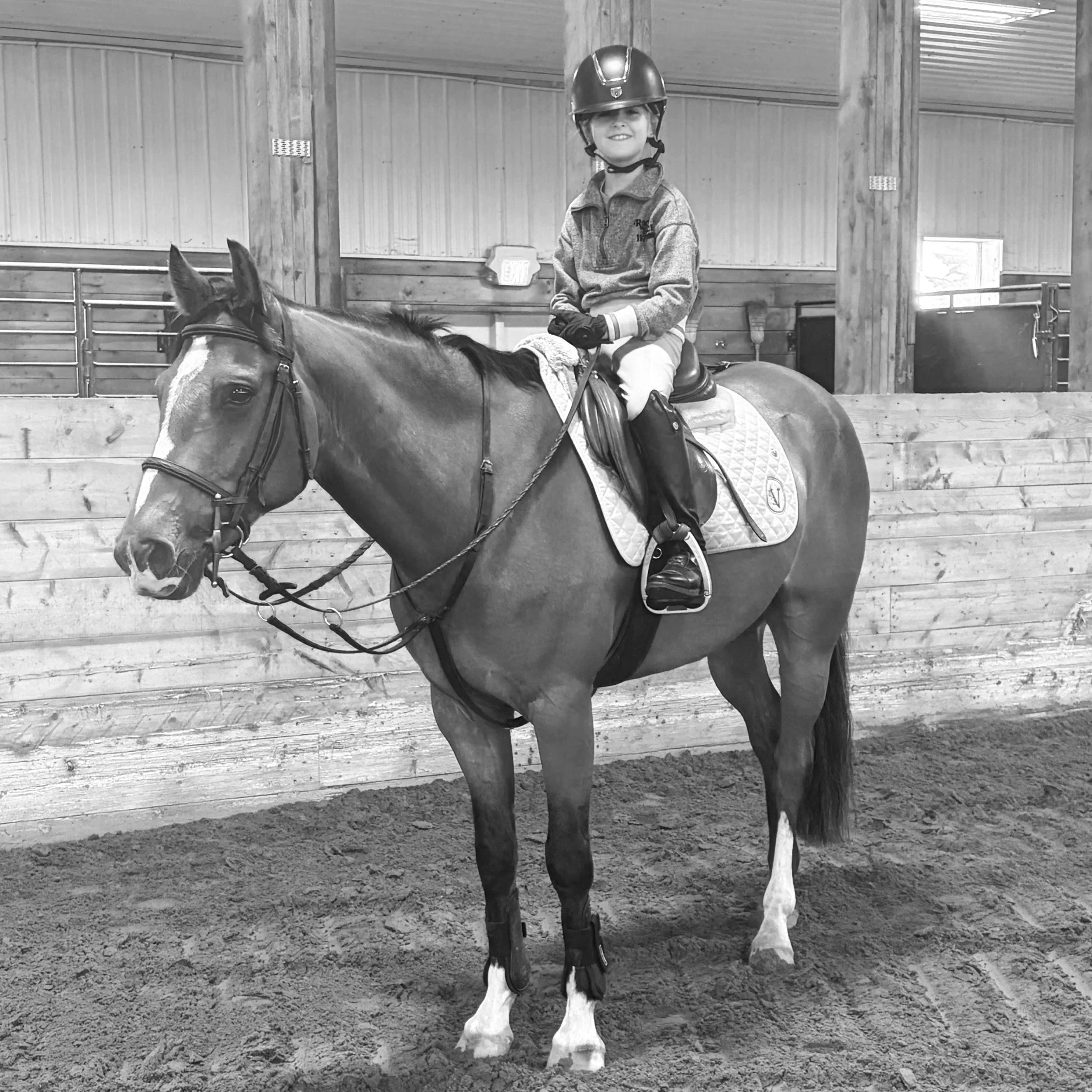 A young girl in riding gear, including a helmet, sitting on a horse in an indoor riding arena with wooden walls.