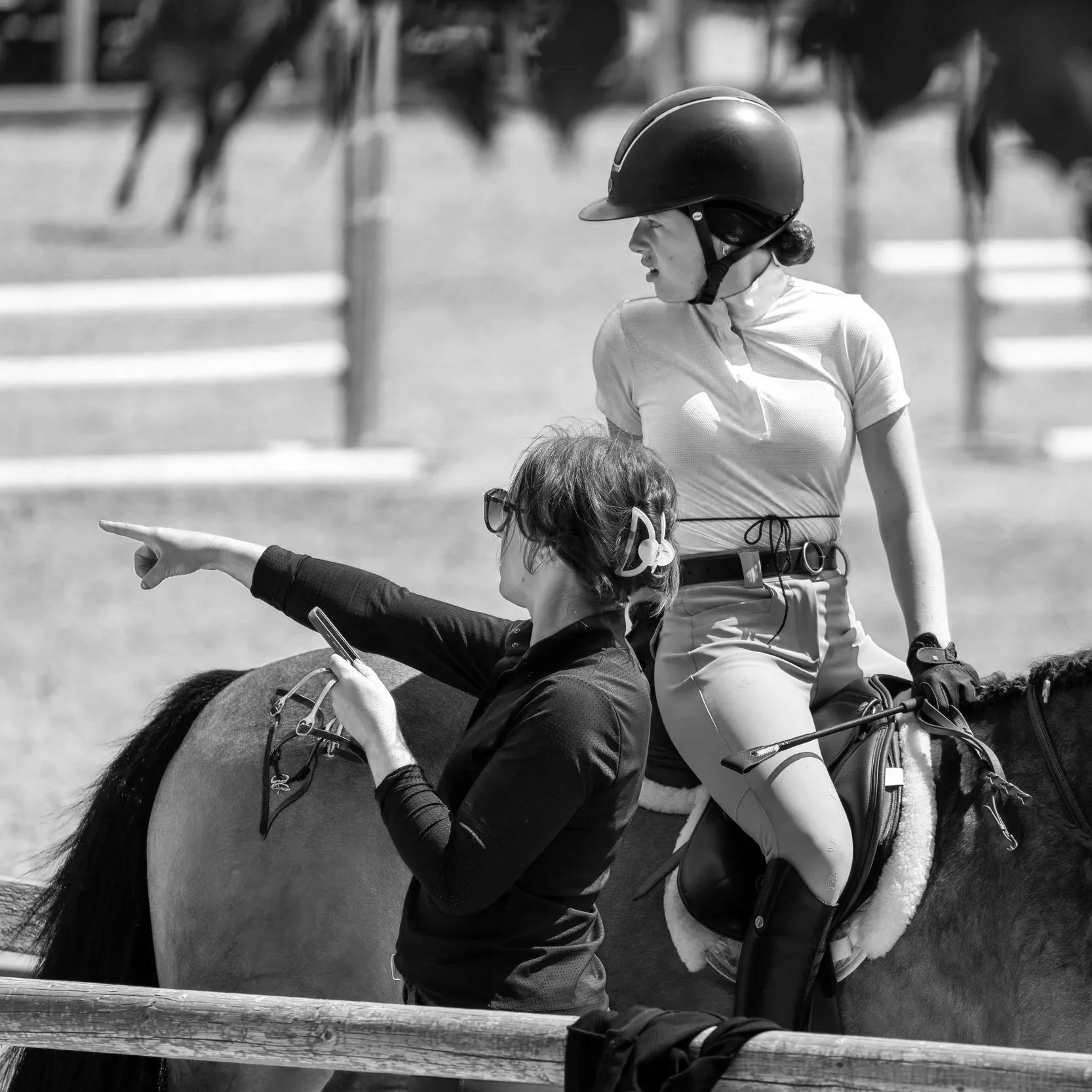 A woman on horseback wearing a helmet and riding outfit looks to her right while a person on the ground points to something. There are horses and fencing in the background.