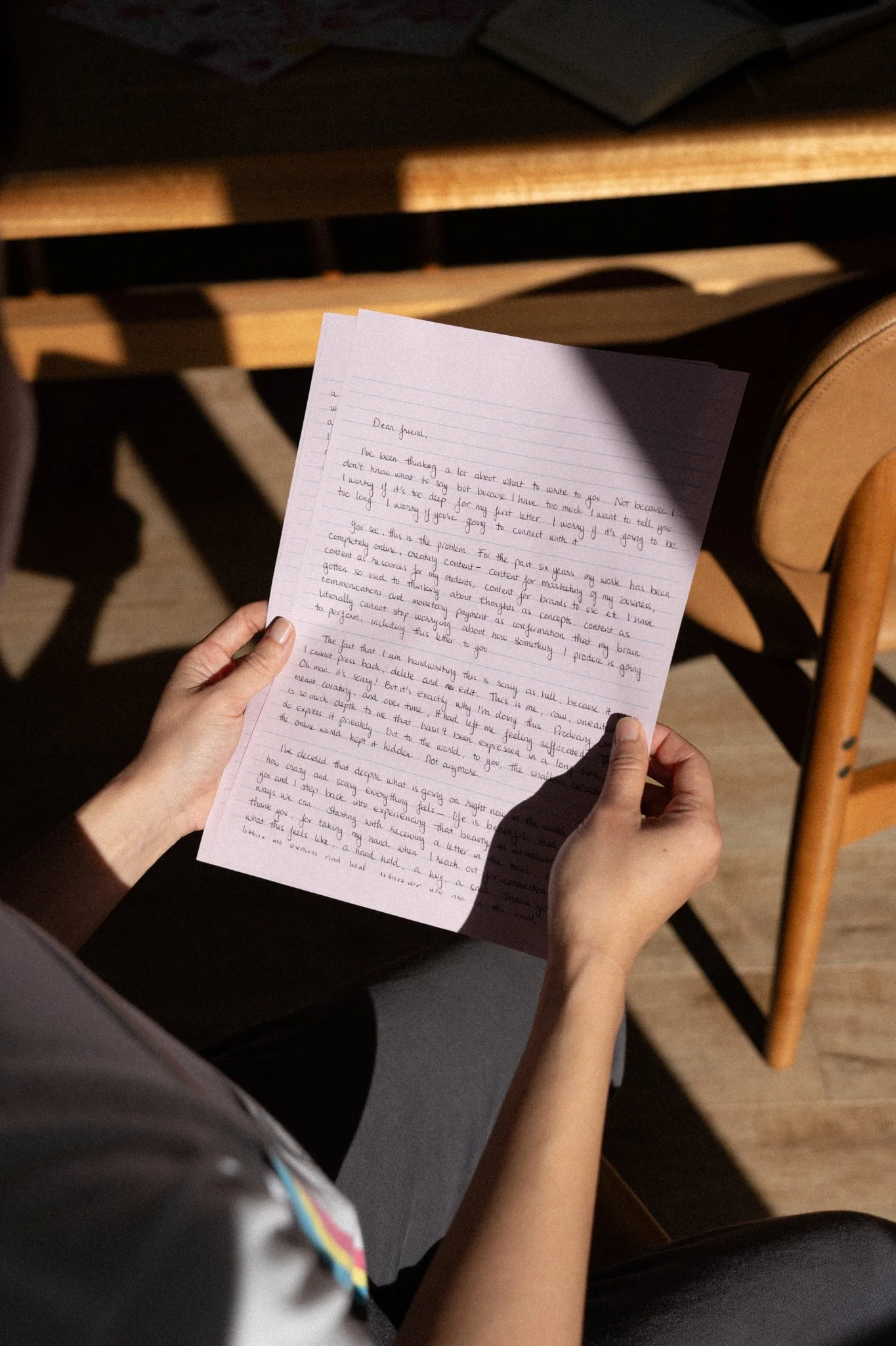 Person holding handwritten letter, seated at a wooden table, with books on top, shadows cast on the letter.