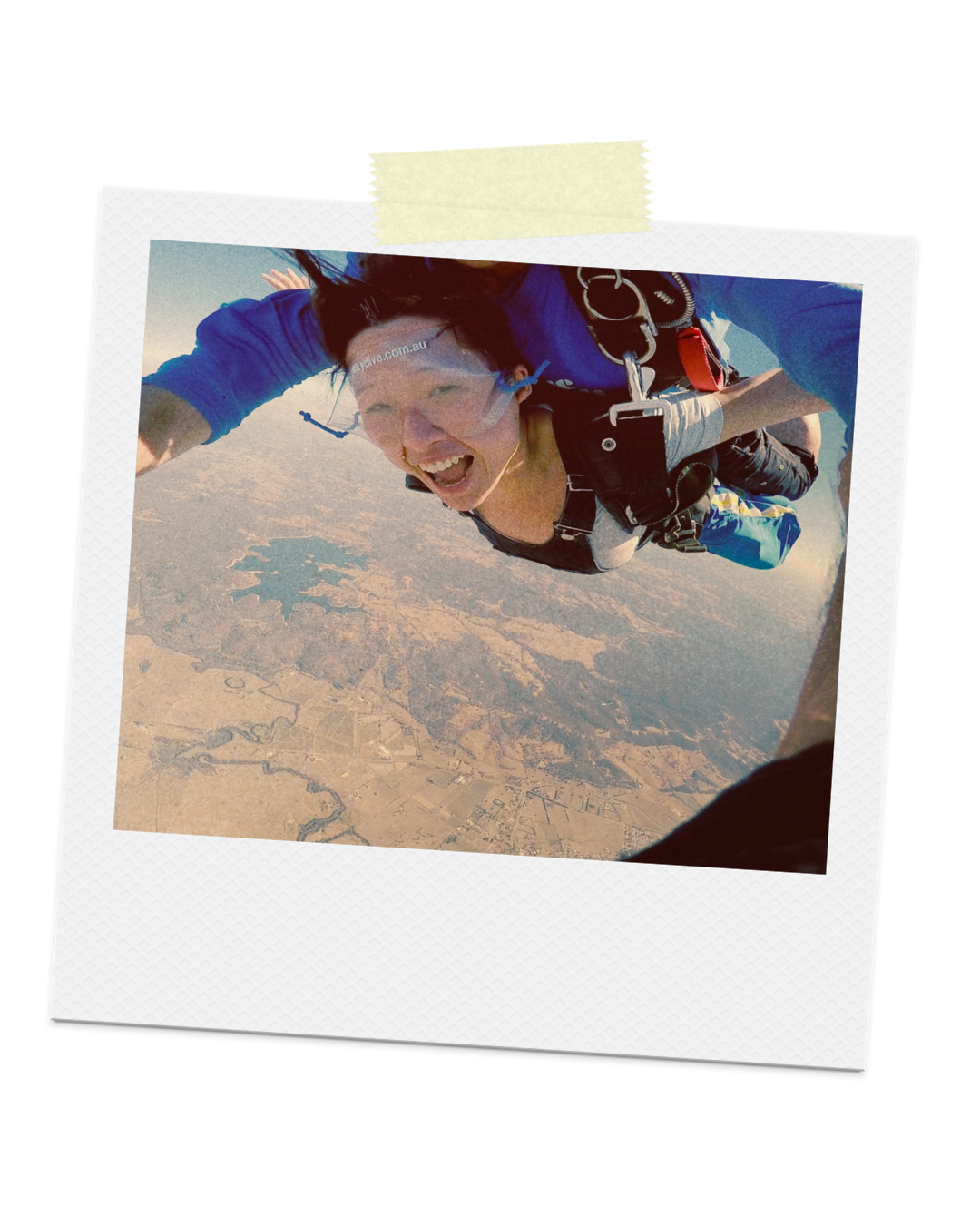 Person skydiving, smiling, wearing goggles and harness, high above the landscape with lakes and fields below.