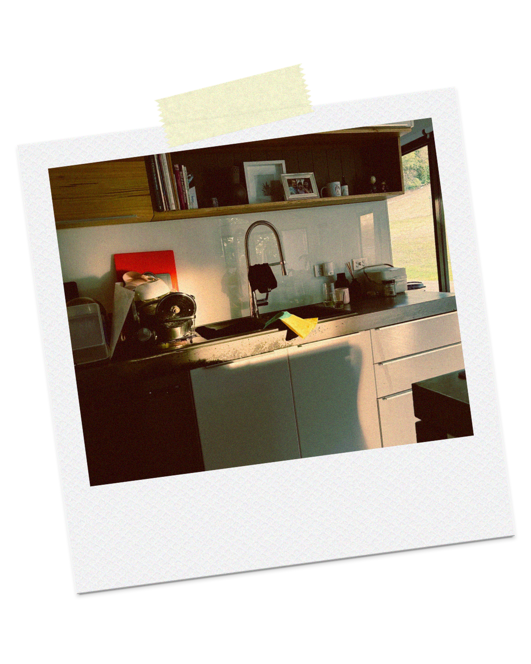 A kitchen with a sink, countertop, shelves, and various kitchen items, with sunlight coming through a window.