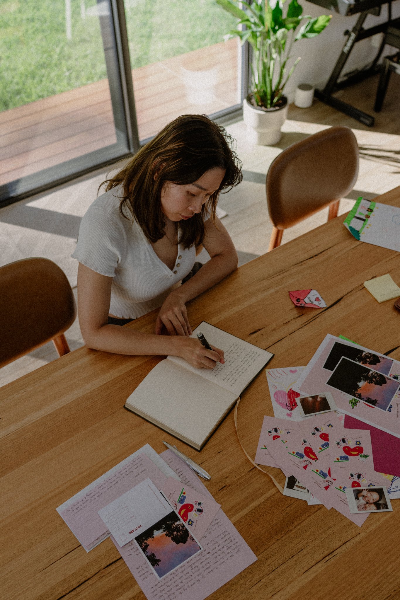 A woman with brown hair wearing a white shirt, writing in a notebook at a wooden table. The table has various papers, colorful drawings, and photographs scattered across it. There is a large window in the background revealing a grassy outdoor area.