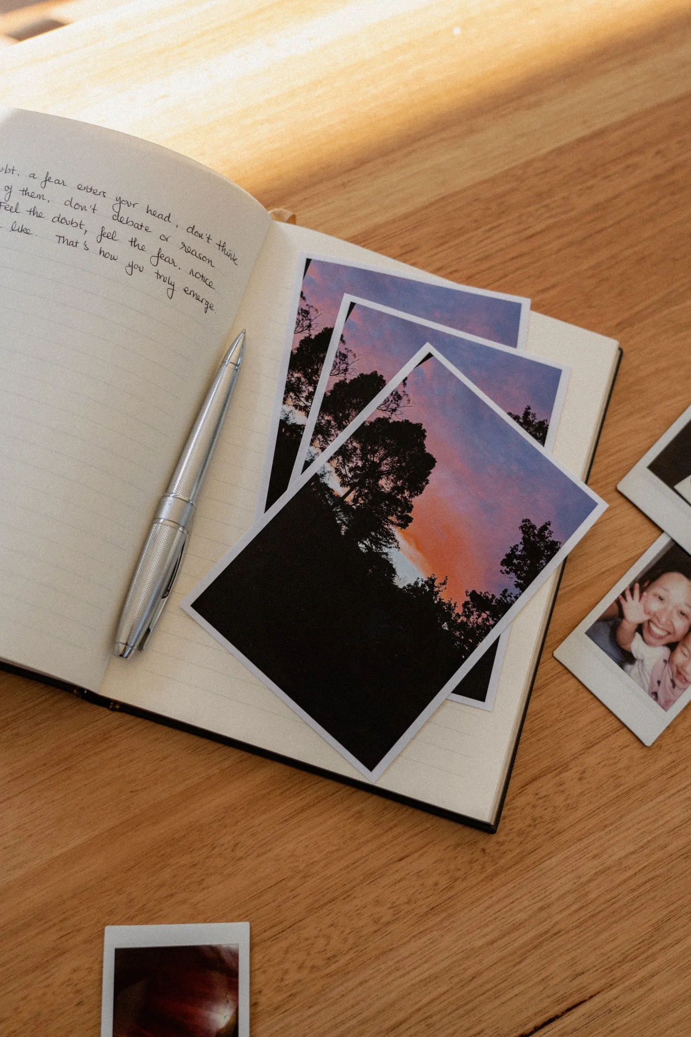 Open planner with handwritten notes and printed sunset photographs on a wooden desk, silver pen placed on the left side of the planner, and various photographs scattered around.