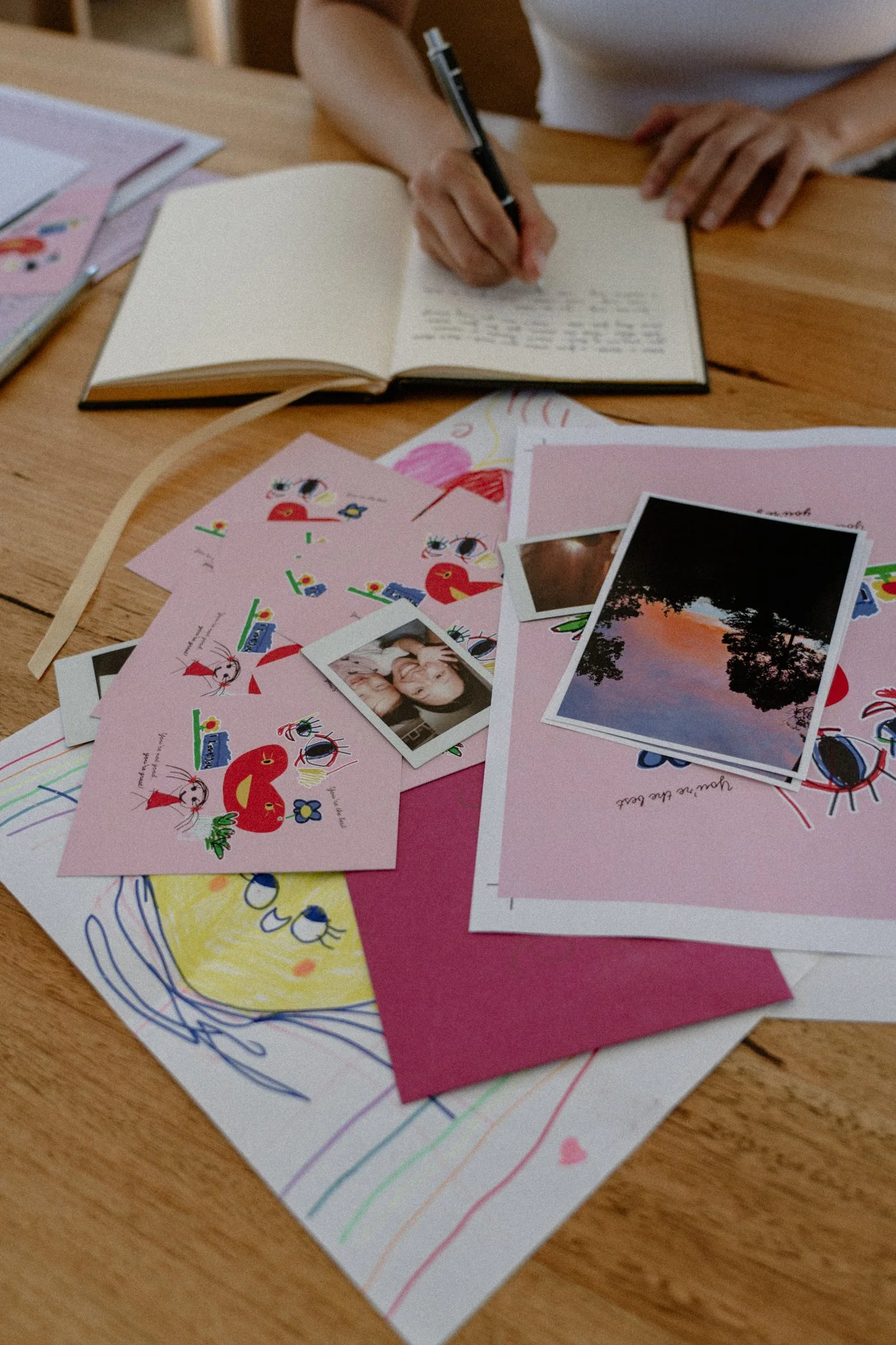 A person writing in a notebook on a wooden table, surrounded by colorful drawings, photographs, and pink and white papers.