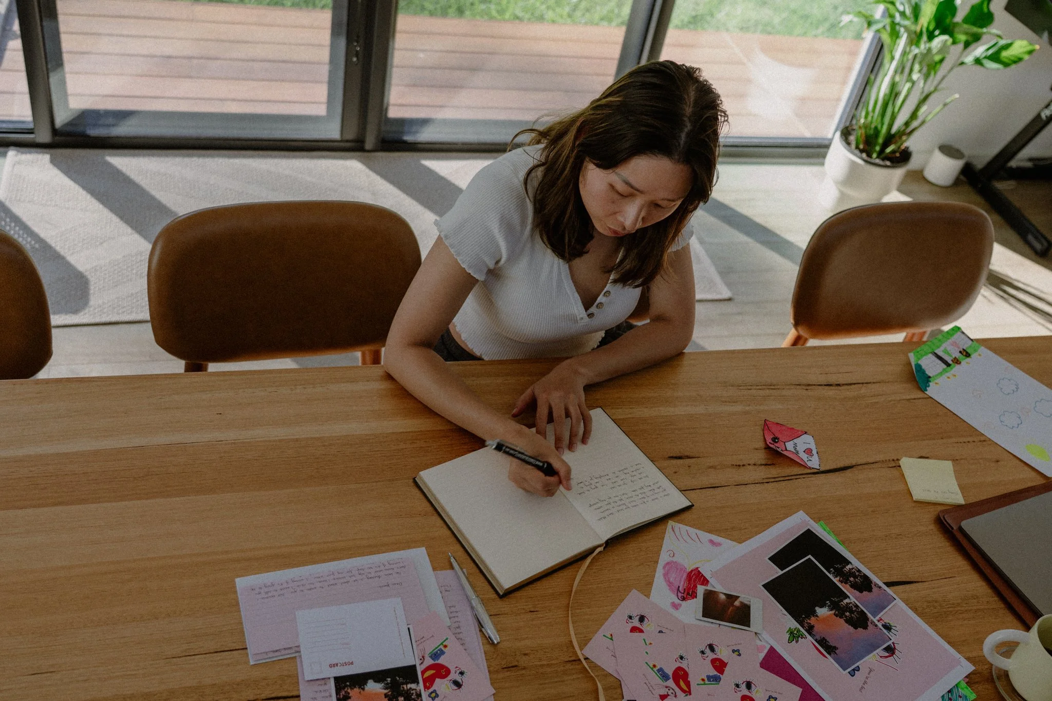 A woman sitting at a wooden table writing in a notebook surrounded by colorful paper crafts, with a large window and sliding glass door behind her showing an outdoor deck and green yard.