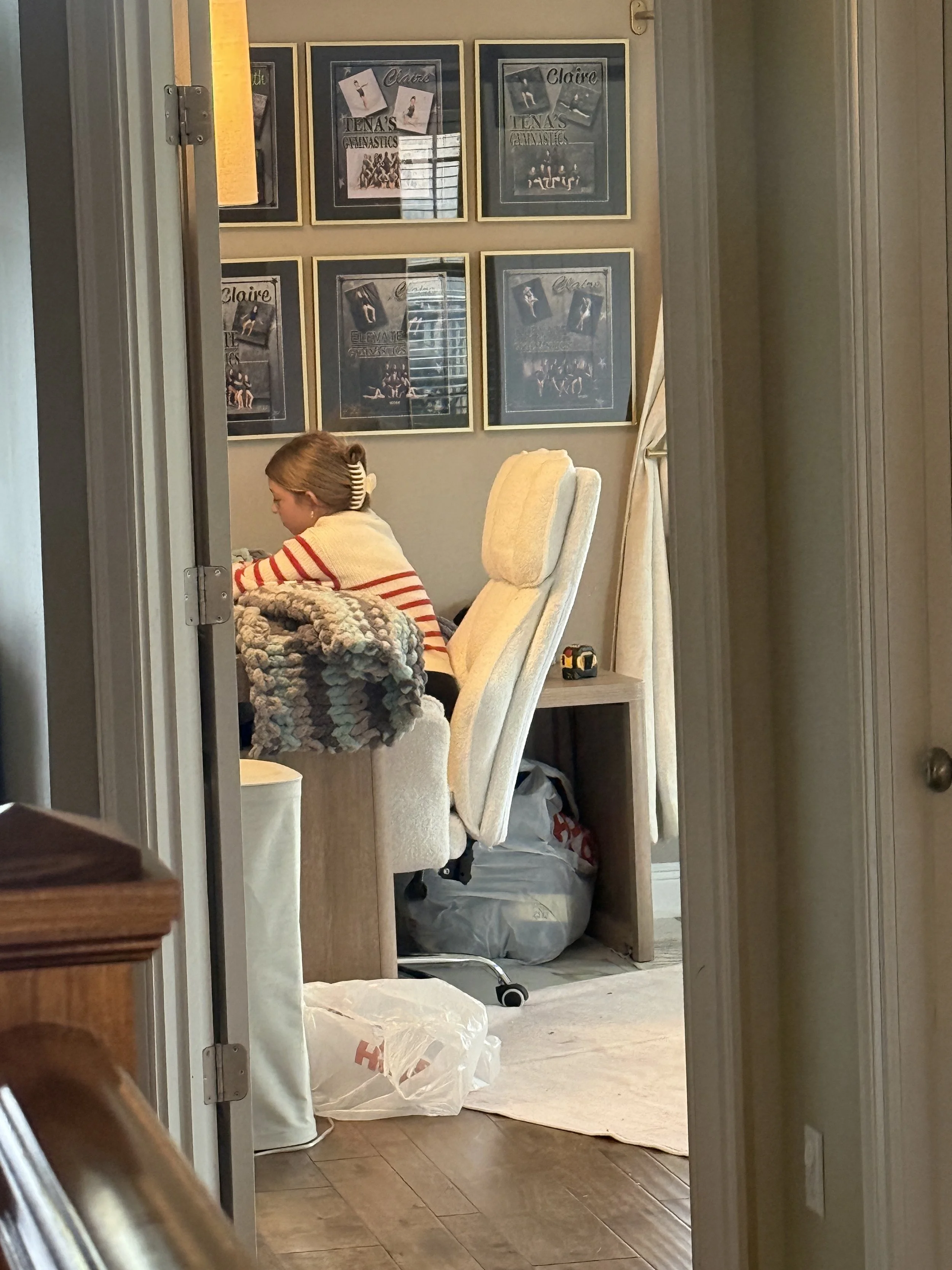 A child sitting at a desk in a cozy room, with framed posters on the wall behind her, a cream-colored chair, and various items on and around the desk.