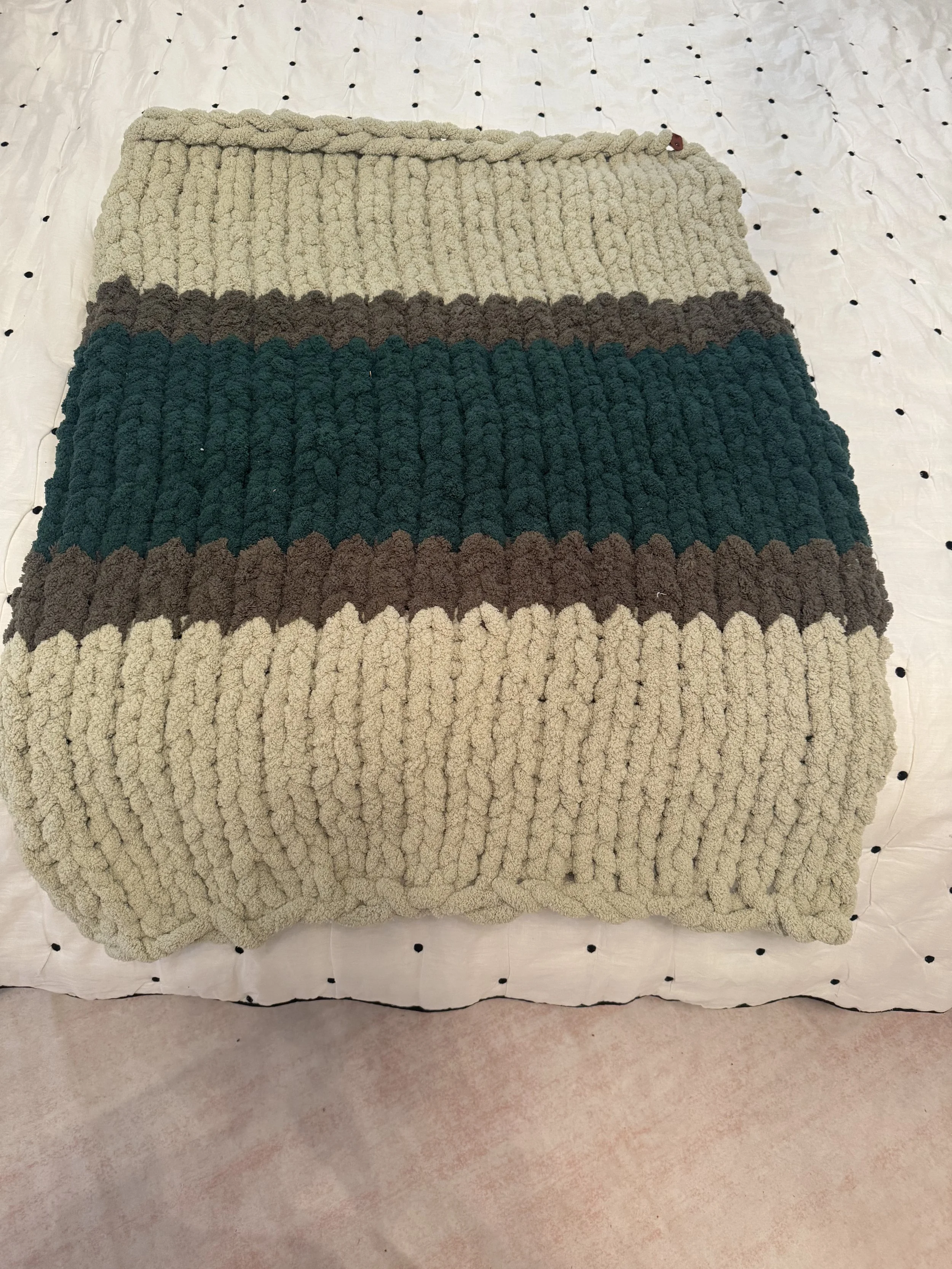 A chunky, striped area rug with beige, dark gray, teal, and brown stripes, laid on a white quilted surface with small black dots.