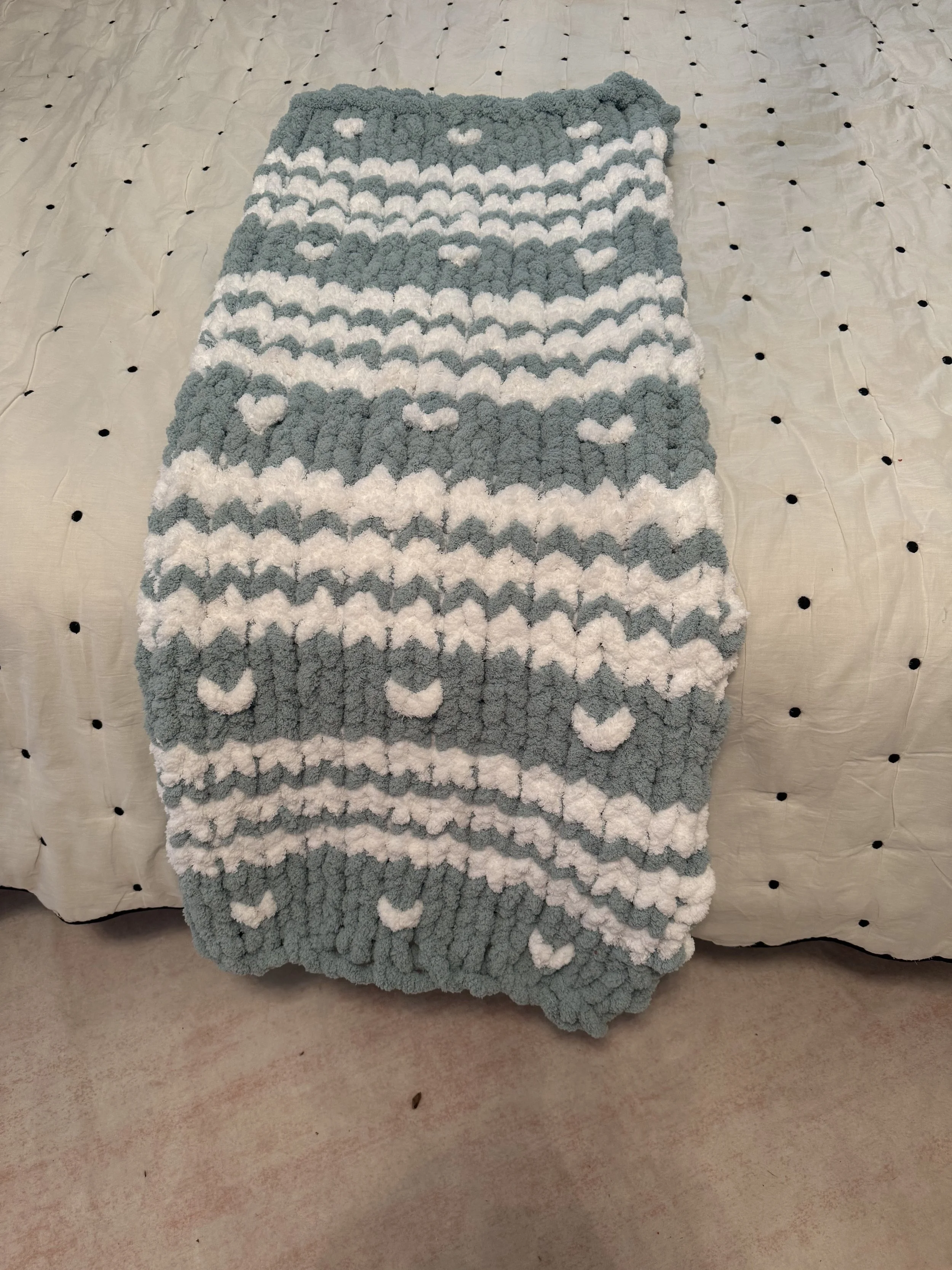 A gray and white fluffy textured bath mat with heart and moose patterns on a bed with a cream-colored quilted bedspread with small black dots.