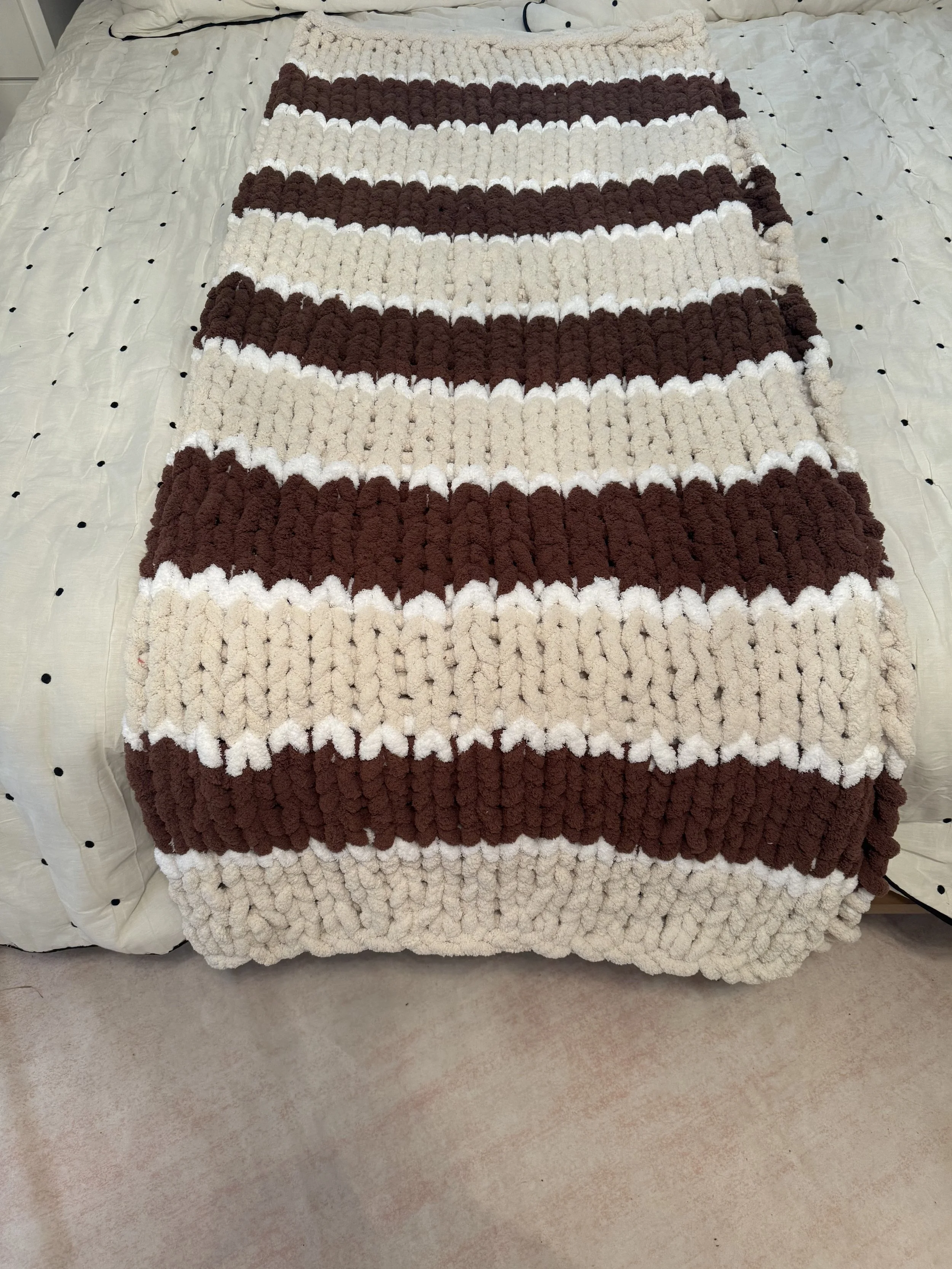 A textured blanket with horizontal stripes in shades of brown, beige, and white, spread out on a bed with a white bedspread and a beige carpeted floor.