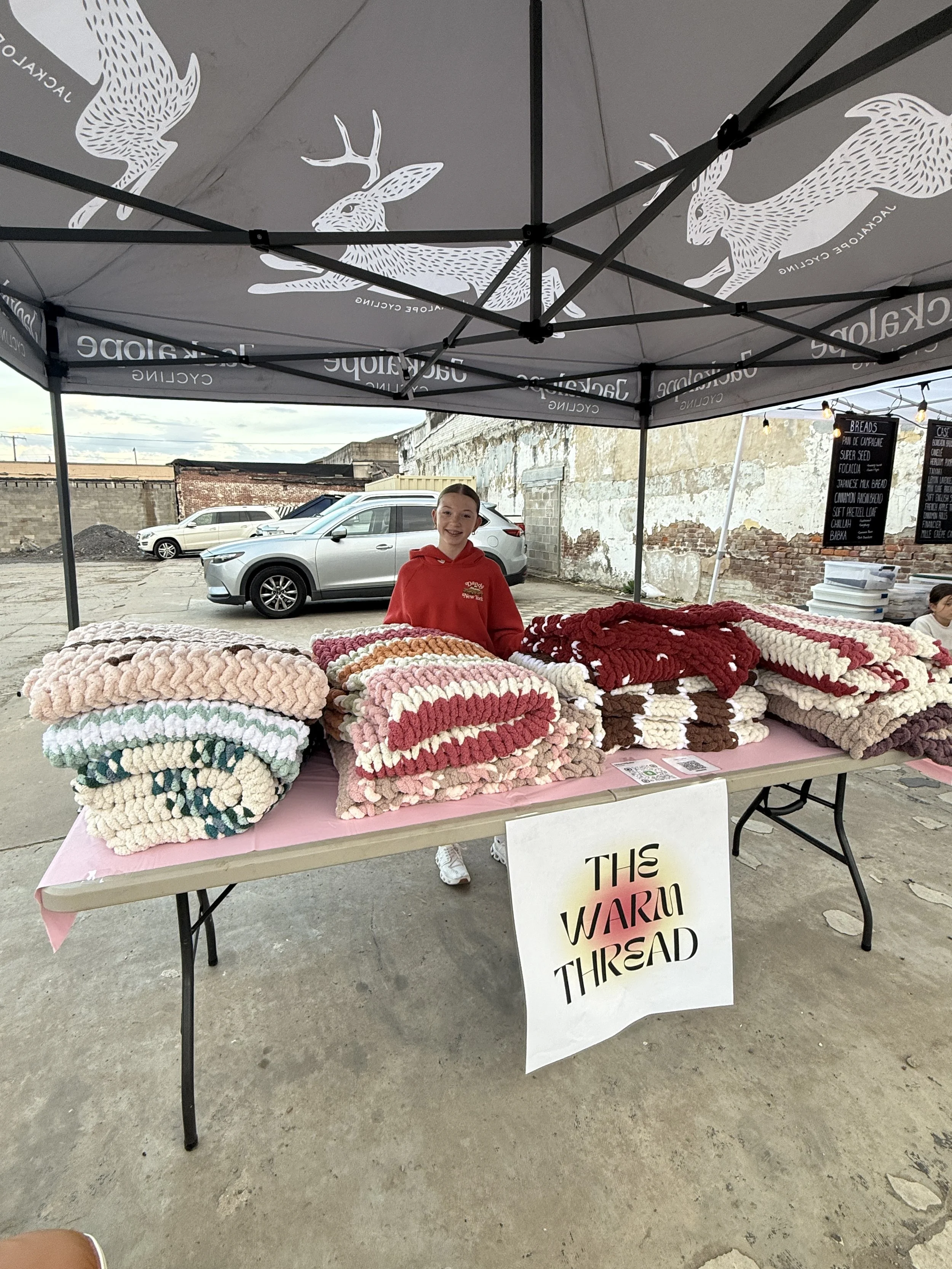 A young girl standing behind a table of cozy, chunky knit blankets at an outdoor market stall, with a sign that reads 'This Warm Thread' hanging in front.
