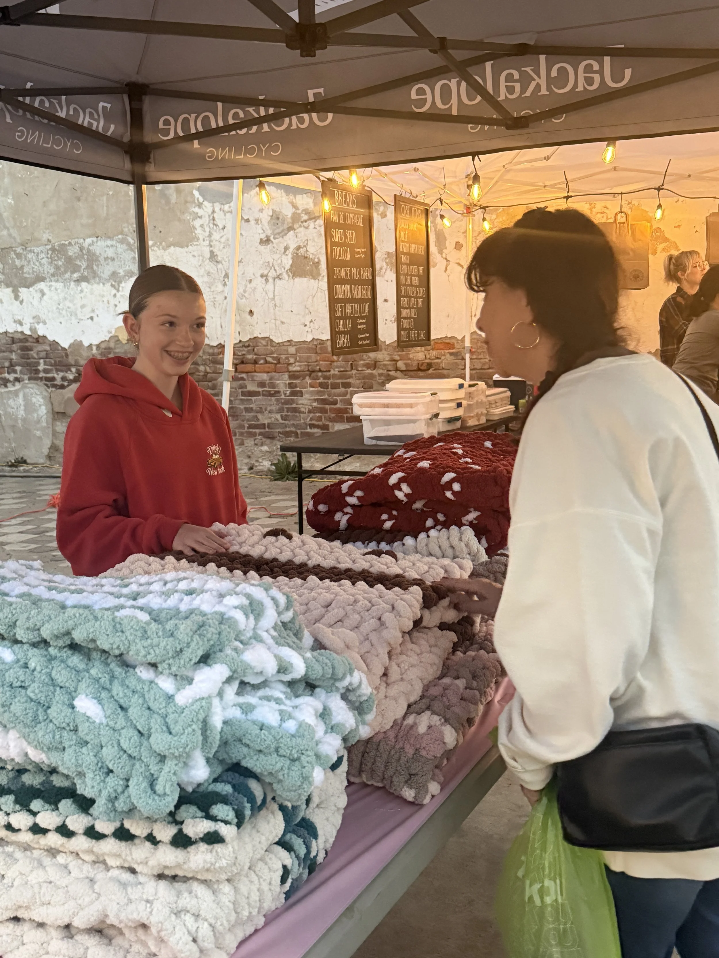 A young woman in a red hoodie is selling colorful, fluffy blankets at an outdoor market stall, engaging with a customer under string lights, against a weathered brick wall.
