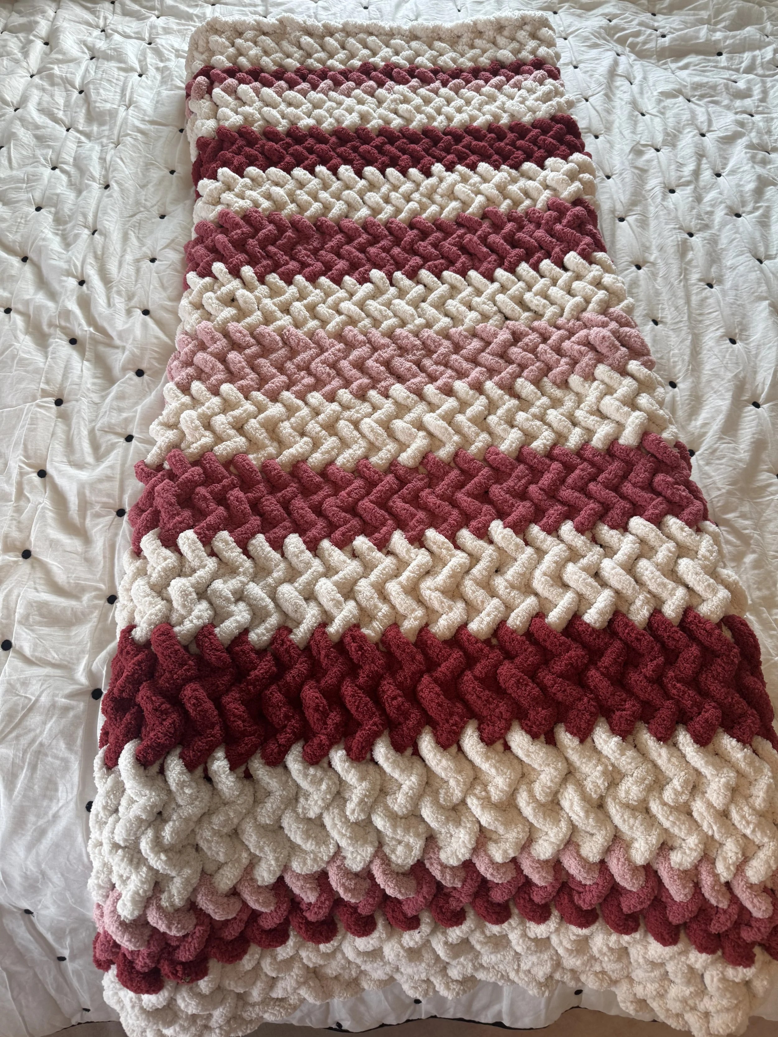 A braided, multi-colored rug with shades of pink, red, cream, and beige, placed on a white quilted bedspread with small black dots.