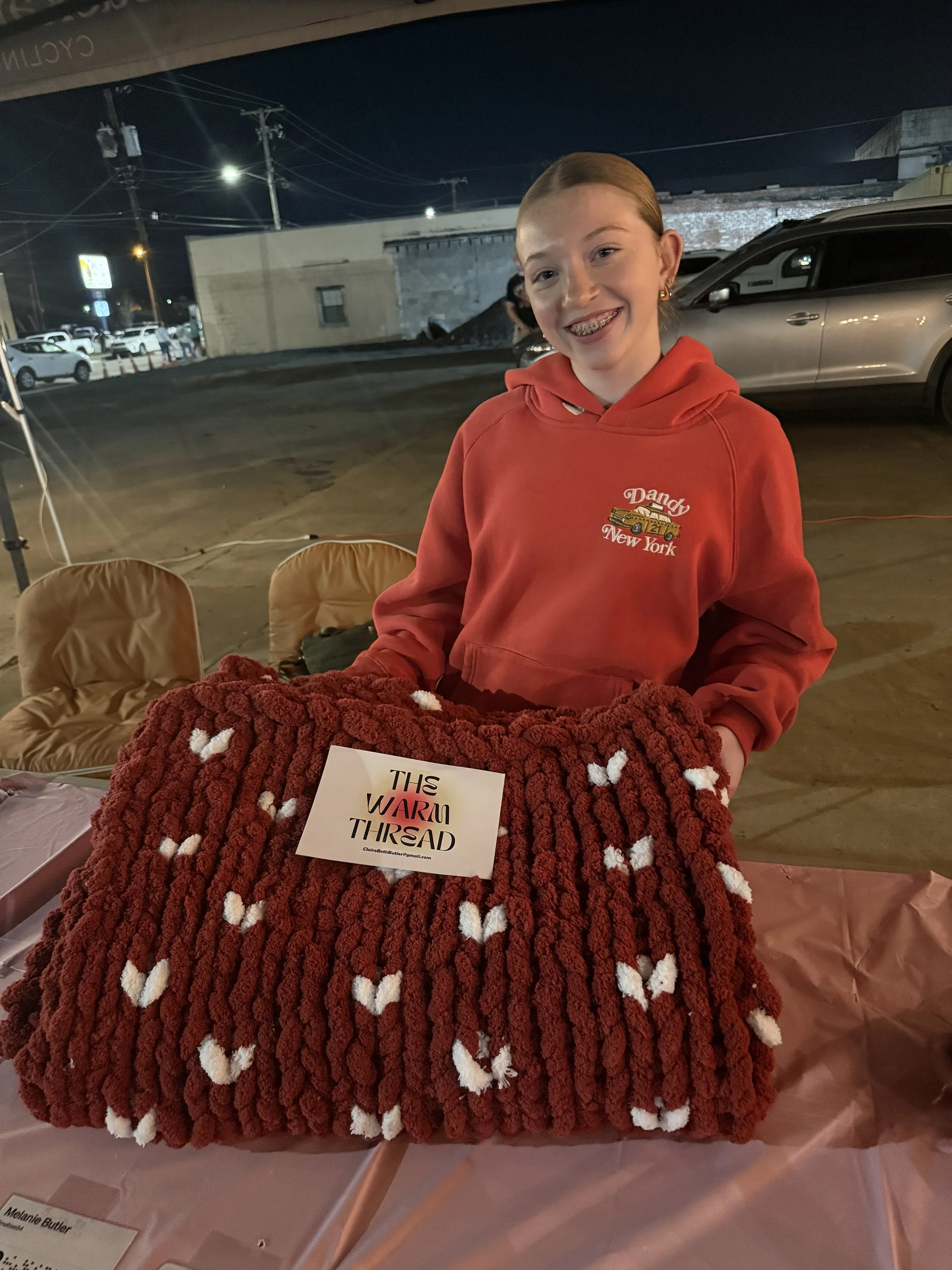 A young girl in a red hoodie with glasses smiling and standing behind a large red and white chunky knit blanket on a table at an outdoor event at night.