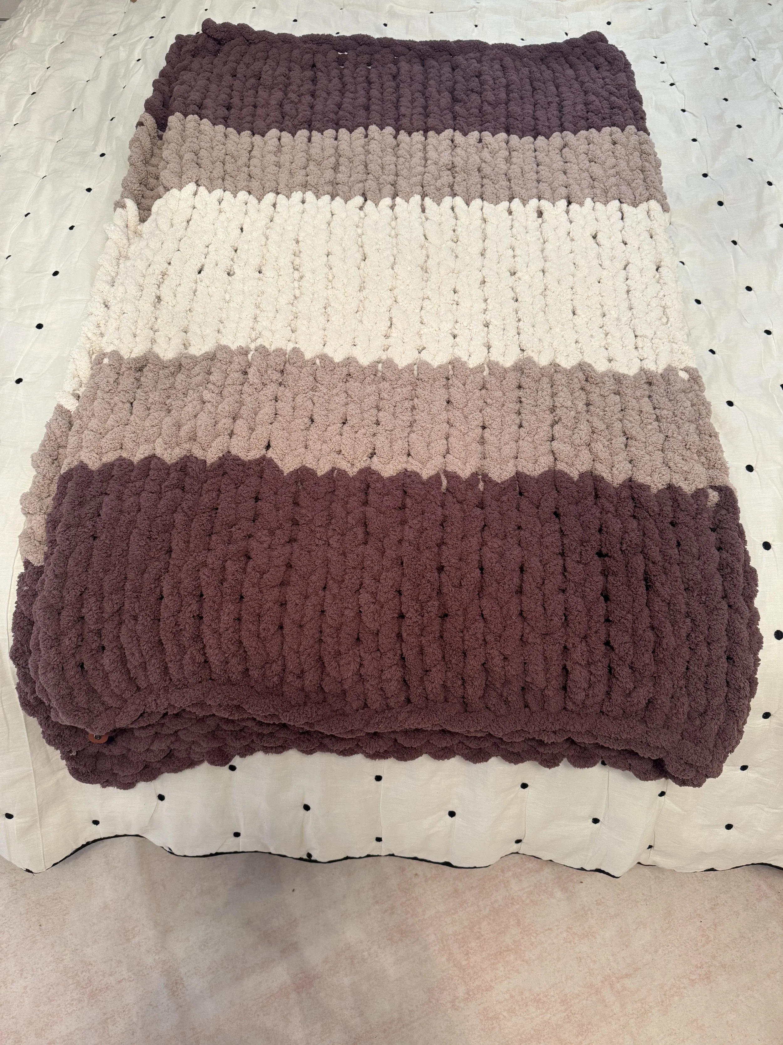 A folded, striped, plush blanket with alternating colors of dark brown, beige, cream, light brown, and maroon, resting on a dotted fabric surface.