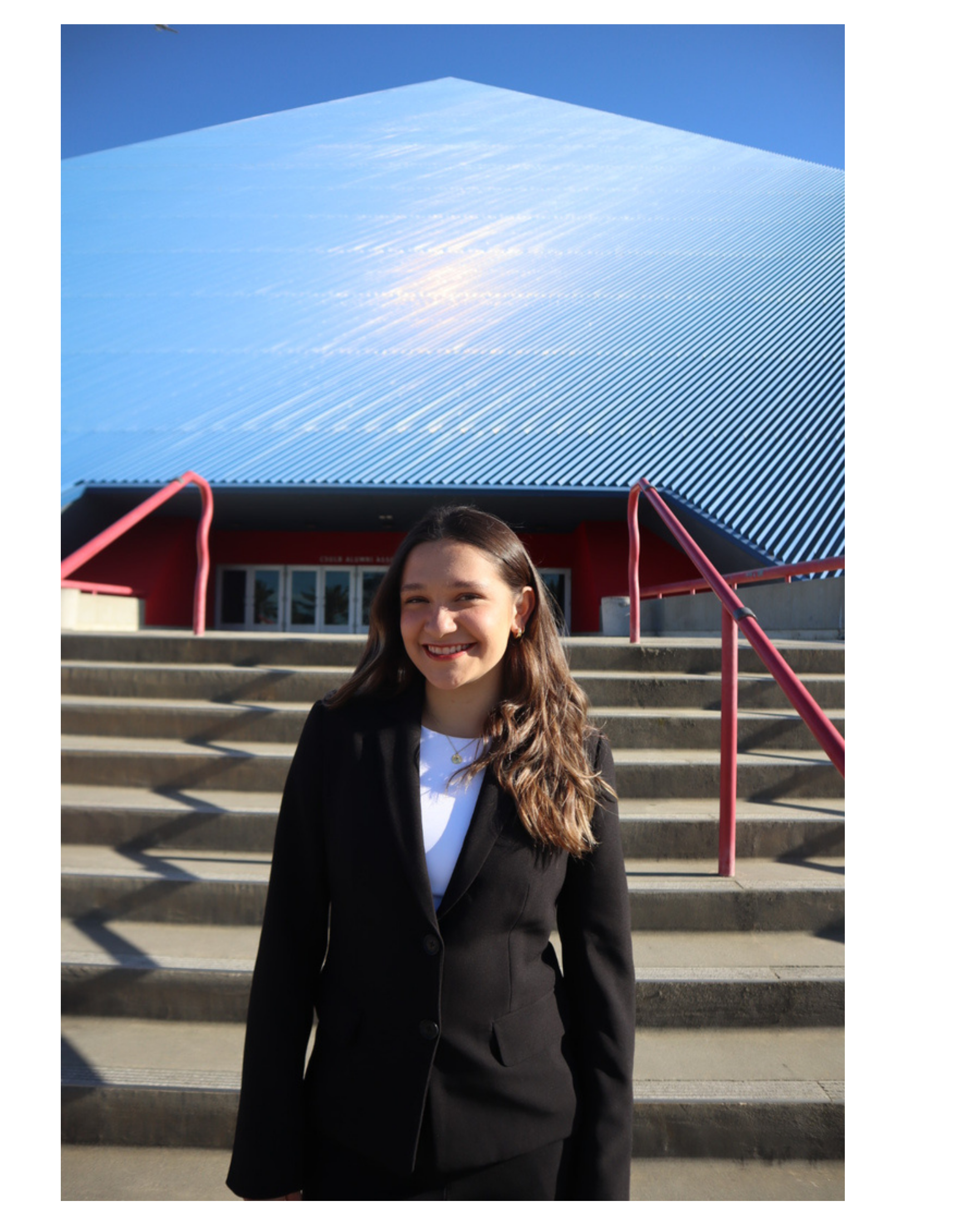 A young woman with long brown hair smiling at the camera, standing on steps in front of a building with a geometric metal facade and blue sky.