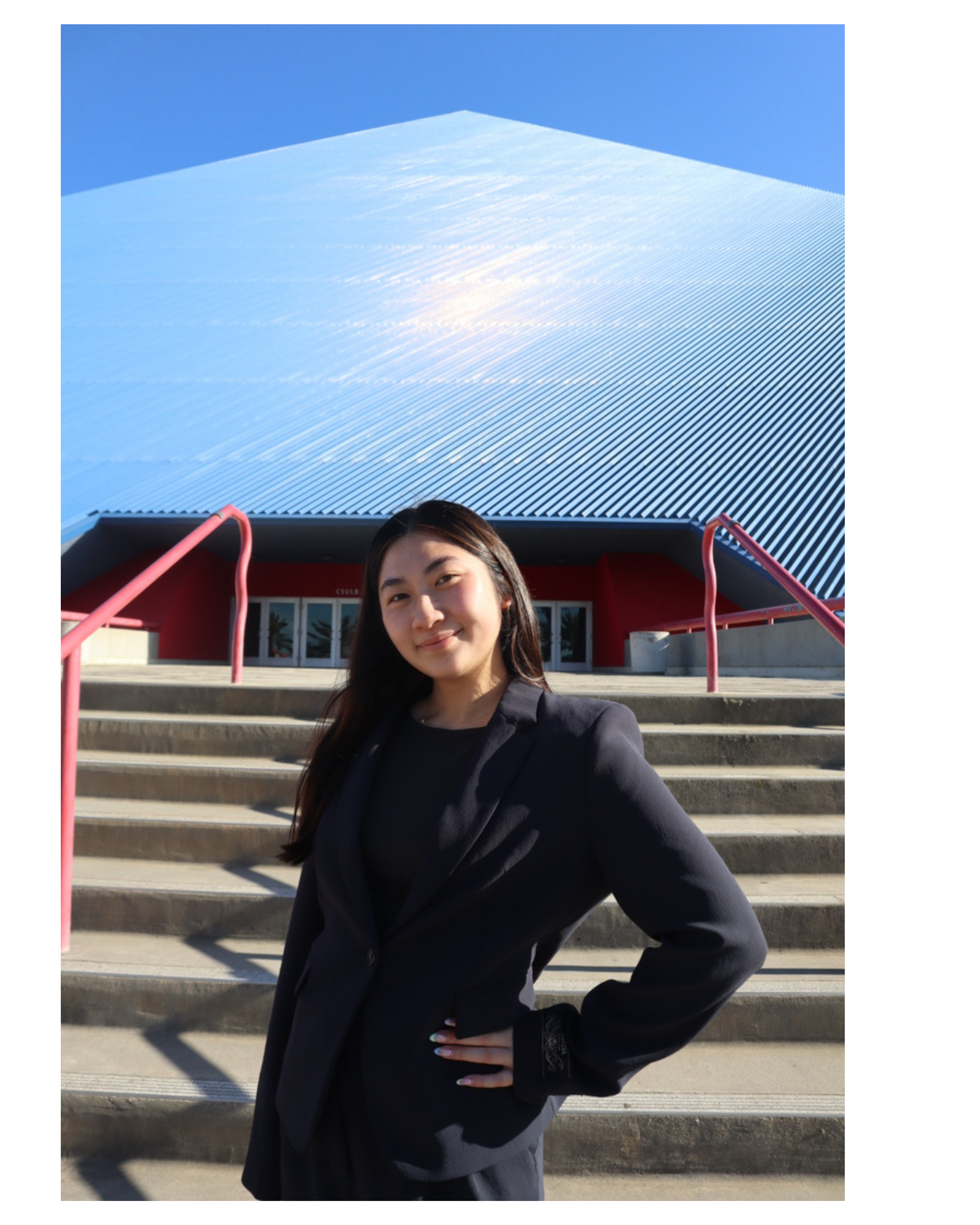A woman with long dark hair wearing a black blazer standing on outdoor steps in front of a building with a modern roof, metal stairs with pink railings, and large glass doors. The sky is clear and blue.
