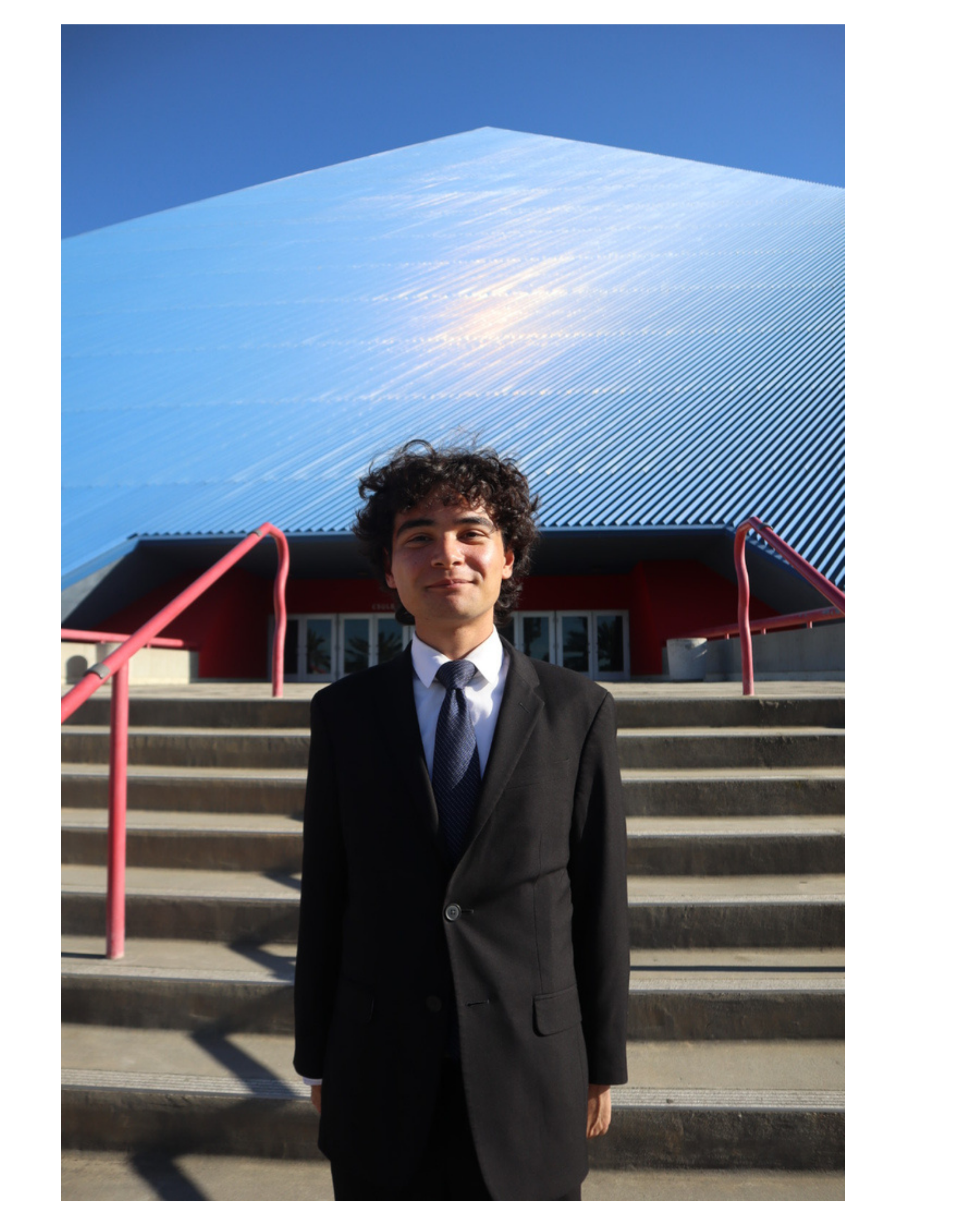 A young man in a black suit and tie stands on external steps in front of a modern building with metallic siding. The sky is clear and blue.