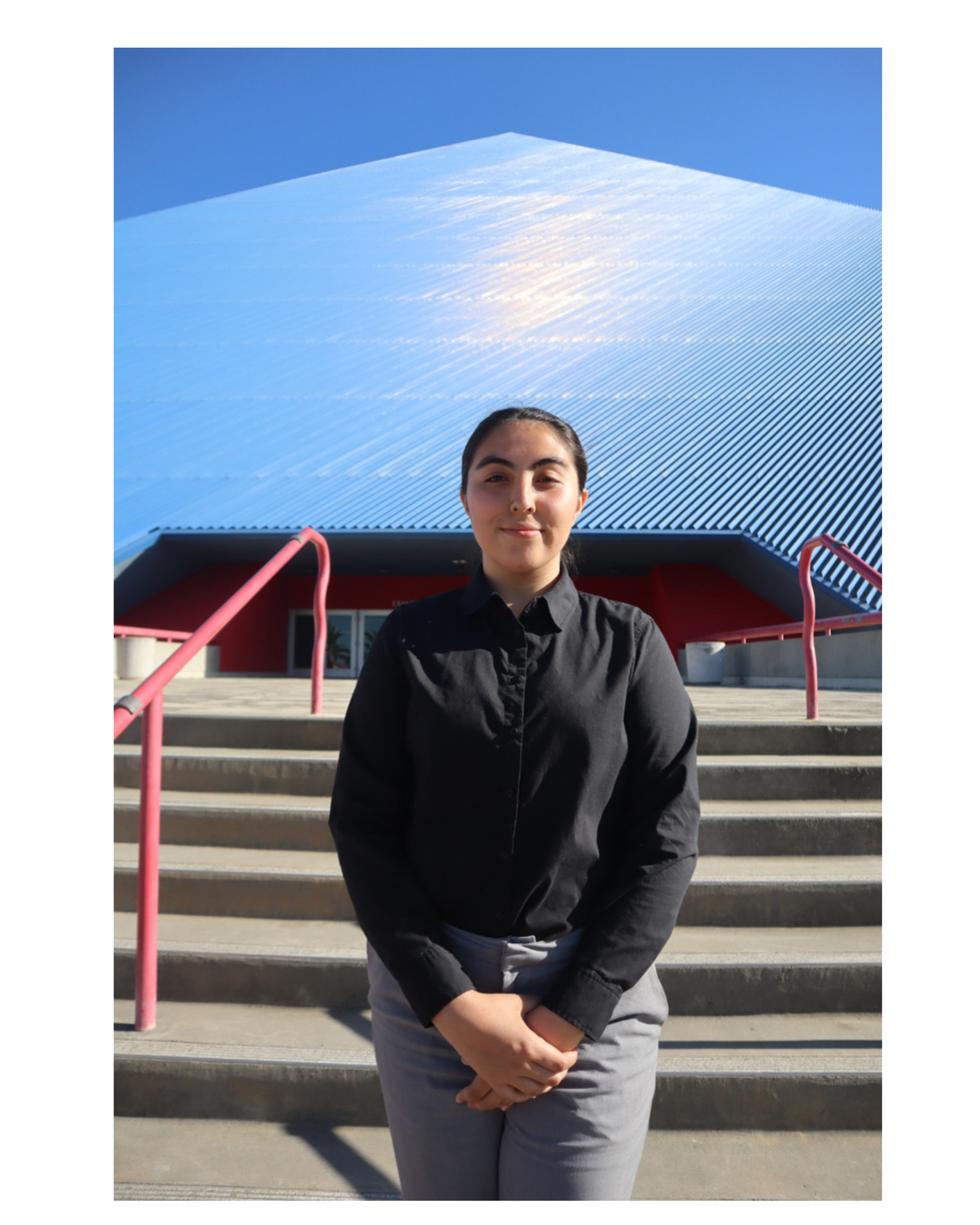 A person with dark hair and light skin standing on stairs outside a building with modern architecture, wearing a black shirt and gray pants, under a clear blue sky.