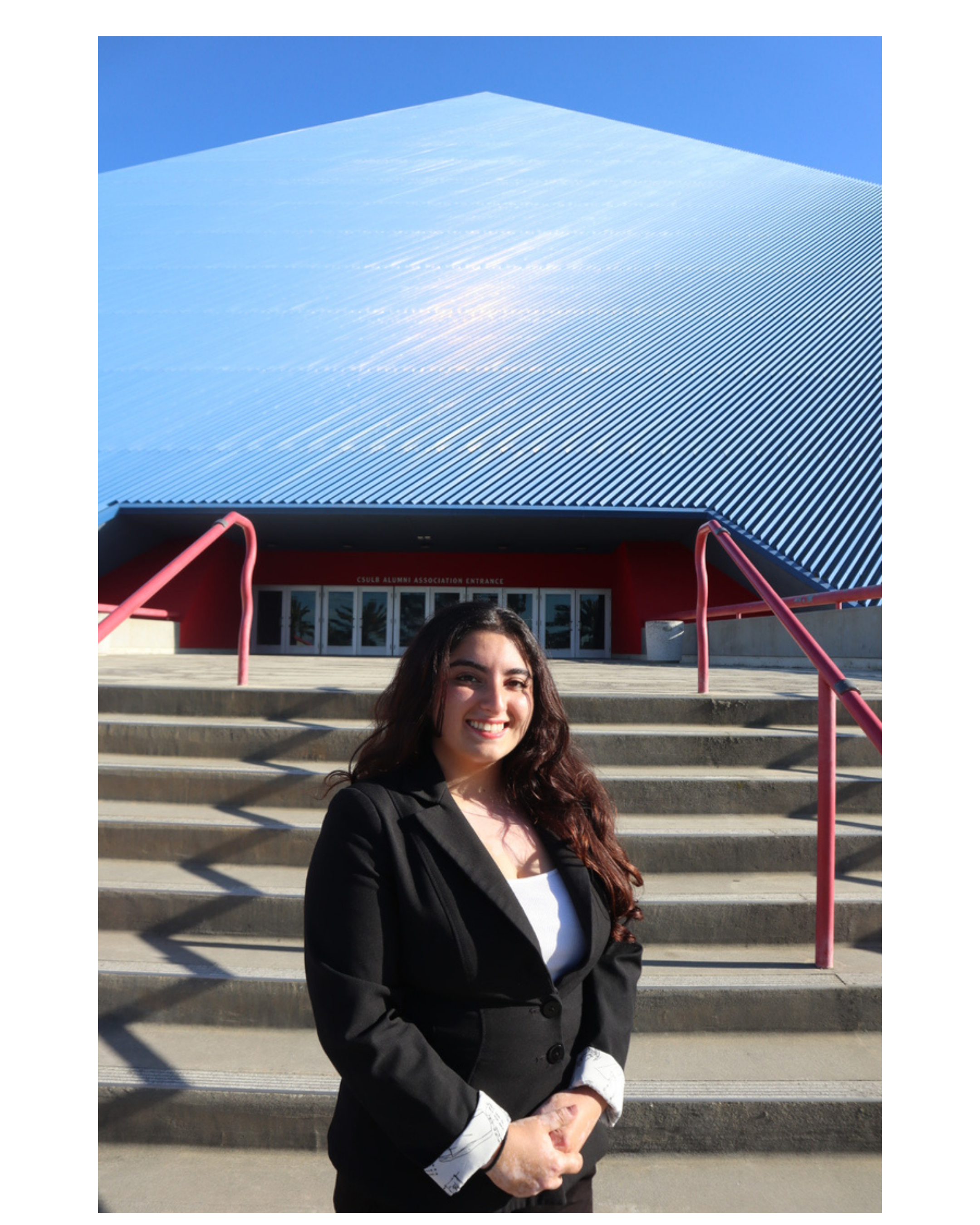 Young woman with long dark hair smiling, dressed in a black blazer and white shirt with patterned cuffs, standing on steps in front of a modern building with a large curved, metallic roof and red stair railings, sunny day.