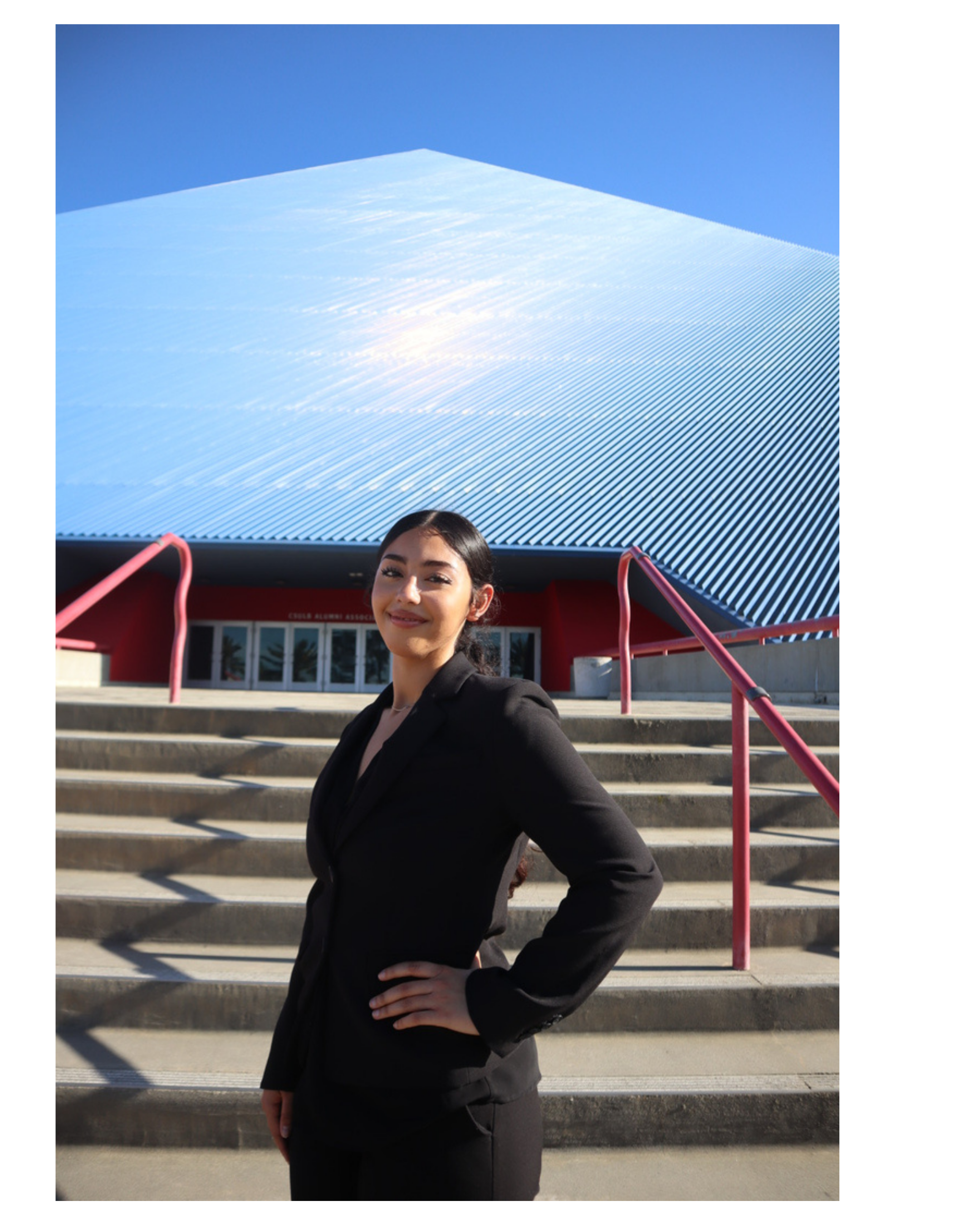 A woman in a black blazer and skirt stands outdoors on steps, with a modern building with a blue ridged roof behind her. She is smiling and has one hand on her hip.