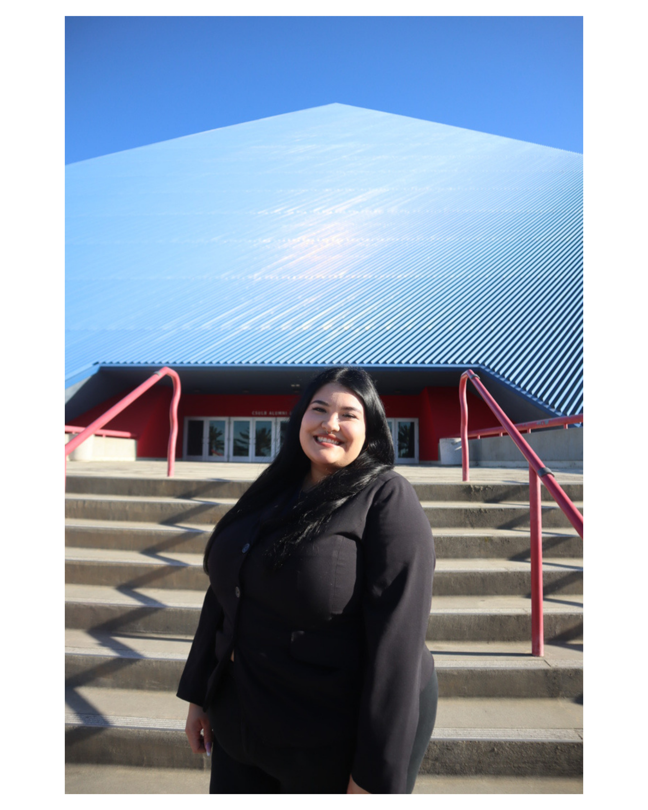 A young woman with long dark hair smiling and standing in front of a building with steps and a metallic, geometric roof structure under a clear blue sky.