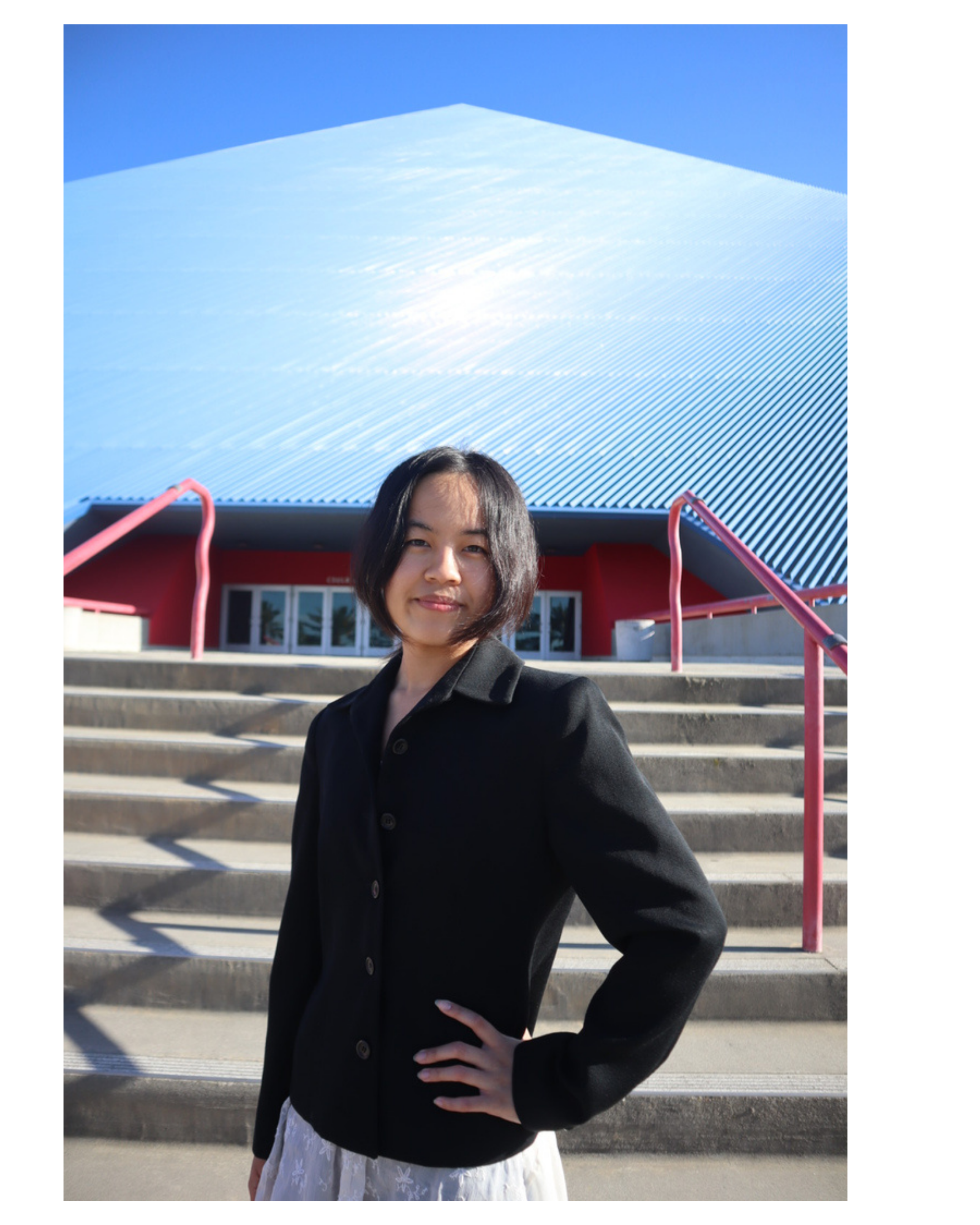 A woman with dark hair, wearing a black jacket and a white dress with floral embroidery, stands on steps in front of a modern building with a metallic roof and red walls, under a clear blue sky.
