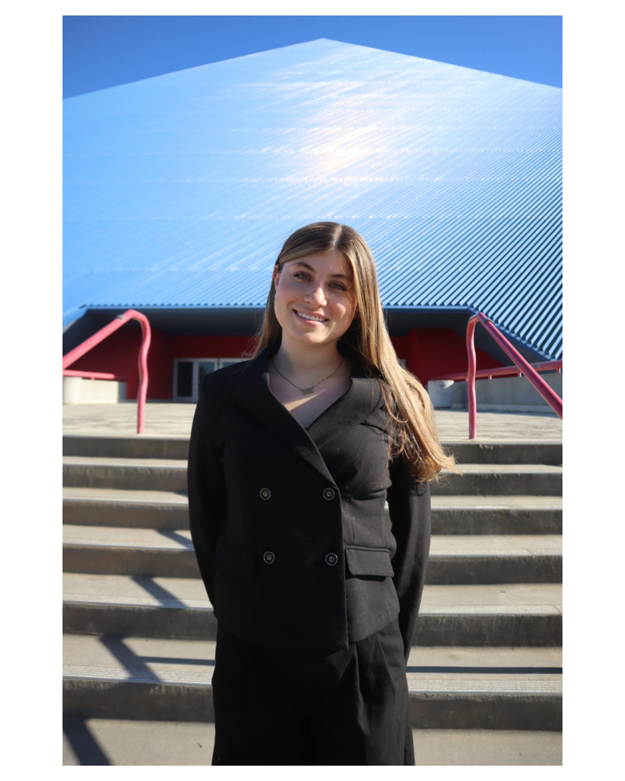 A smiling young woman with long blonde hair standing outside in front of stairs, with a modern building with a blue sky in the background.