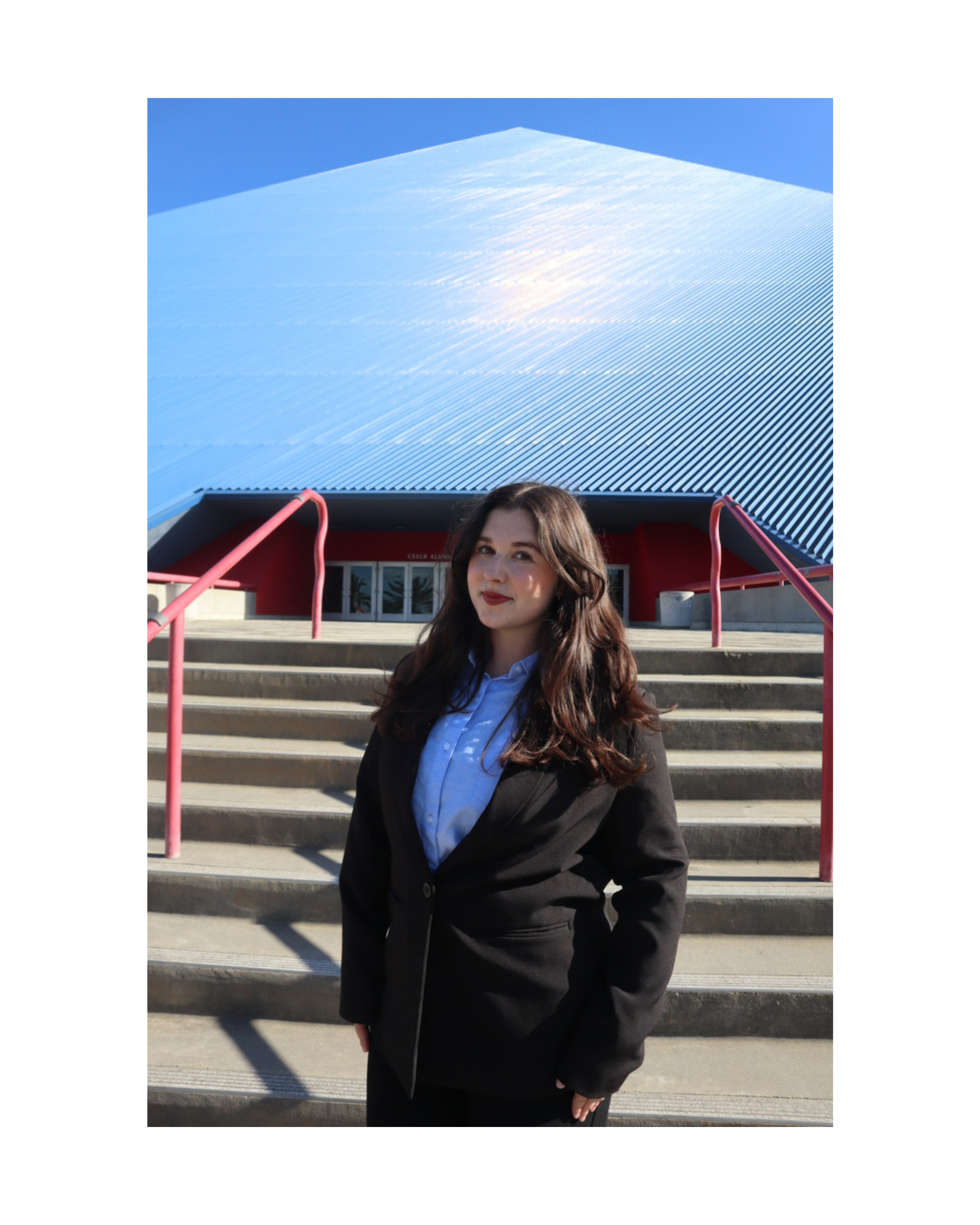 A woman with long brown hair, wearing a black blazer, white shirt, and dark pants, standing on outdoor stairs in front of a modern building with a gray and blue facade. The sky is clear and blue.