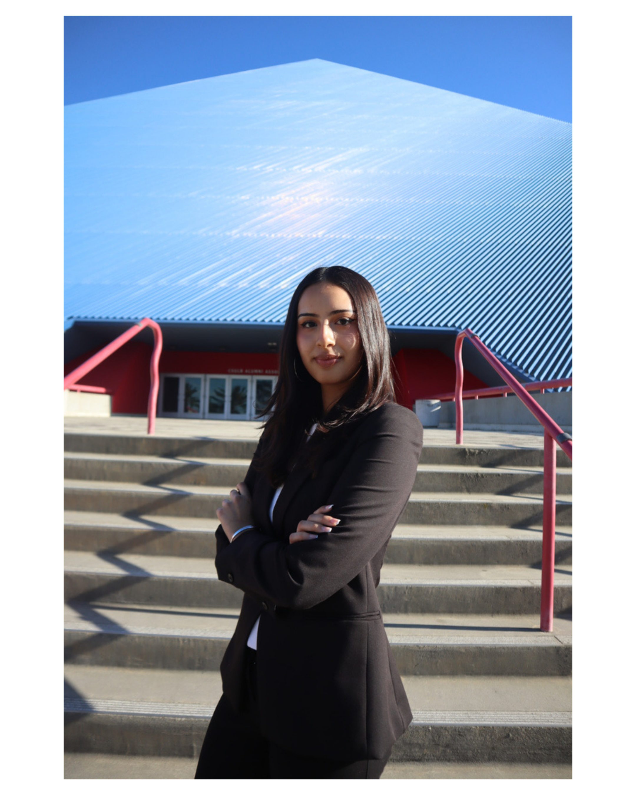 A young woman with long dark hair wearing a black blazer standing outdoors on concrete stairs with red railings, with the Long Beach State Pyramid in the background.