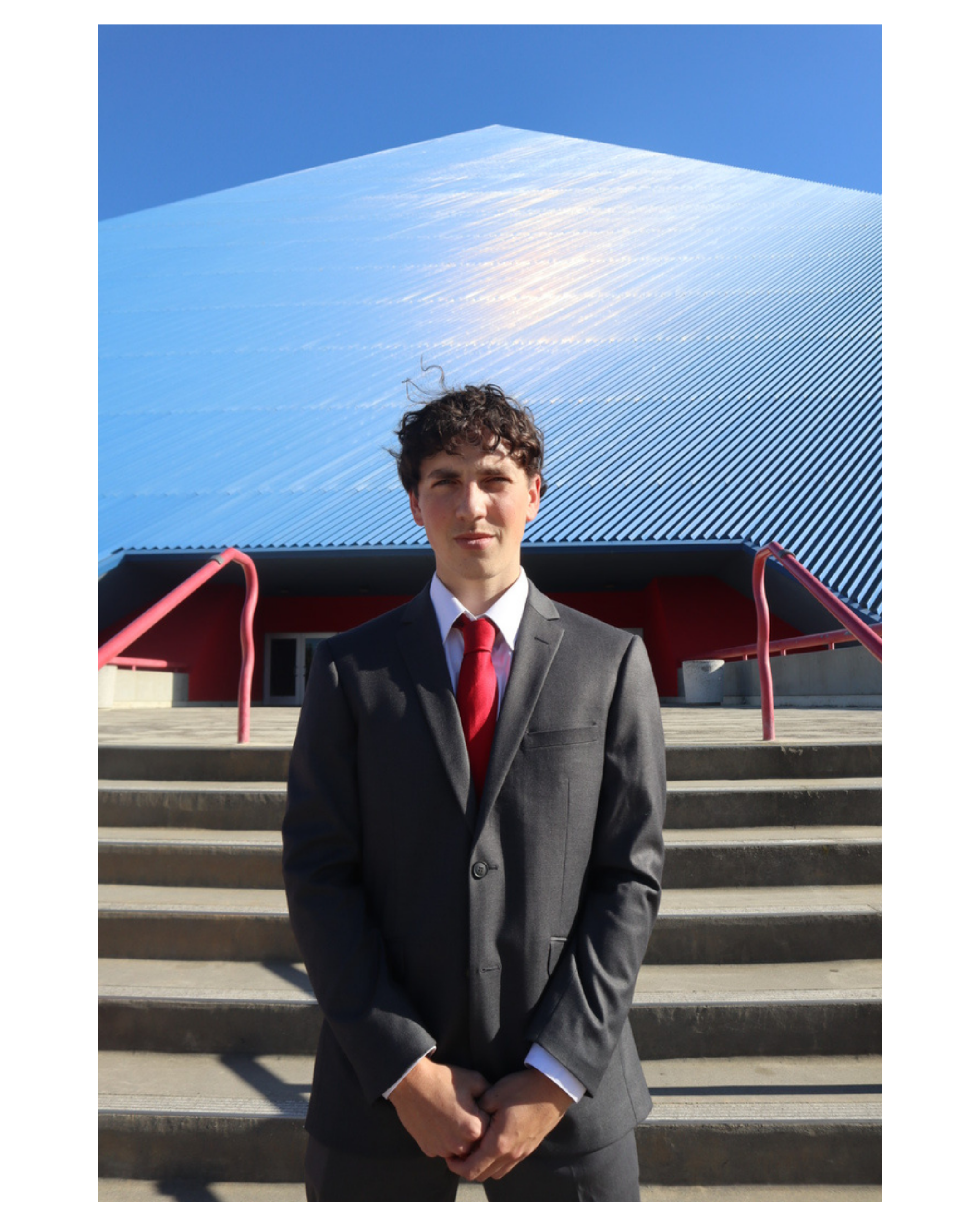 A young man with curly hair dressed in a dark suit, white shirt, and red tie standing on steps outside a building with the Long Beach Pyramid, holding his hands in front of him and looking into the camera.