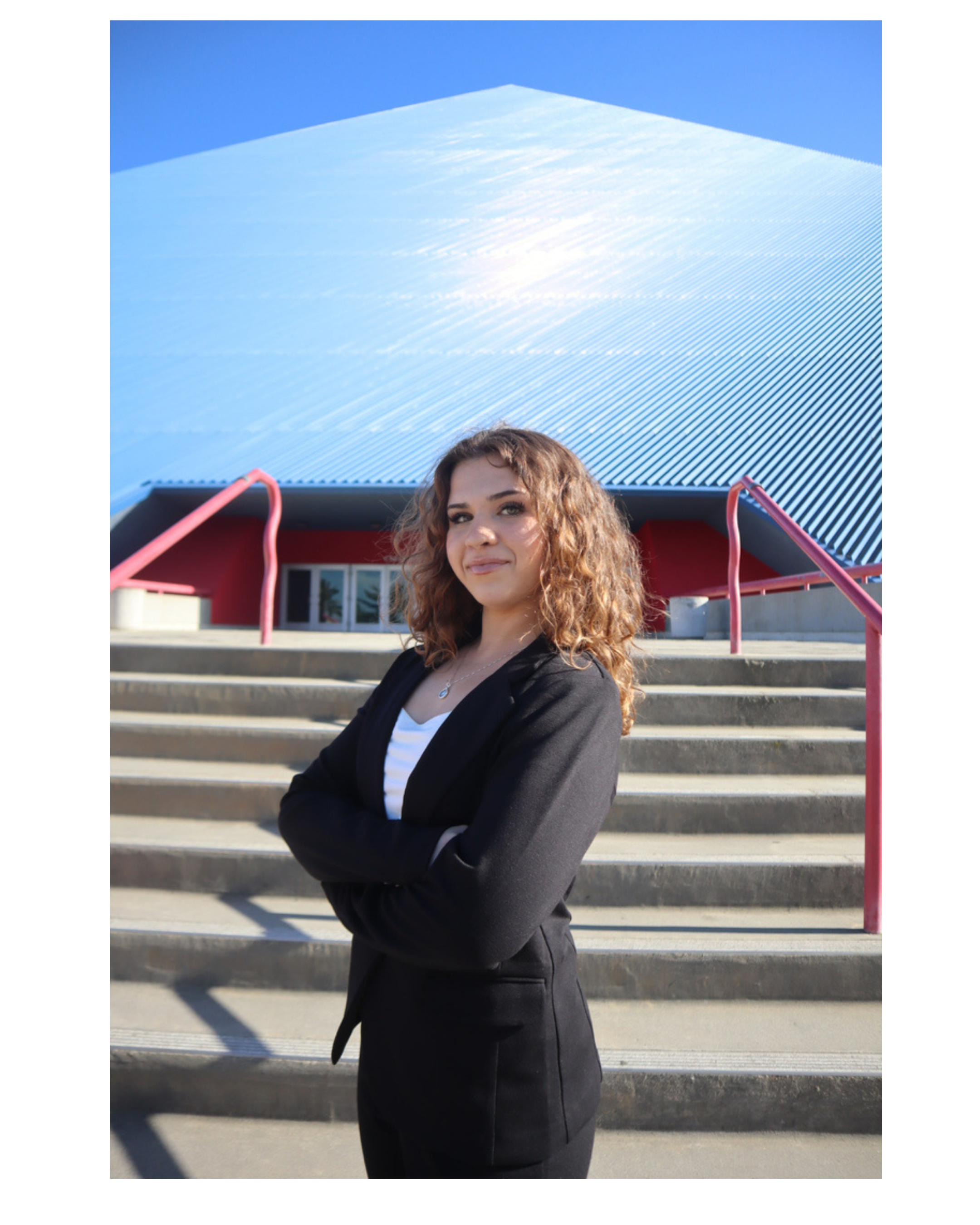 Young woman with curly brown hair standing outdoors on steps, wearing a black blazer and smiling, with the LBSU Pyramid and red accents in the background.