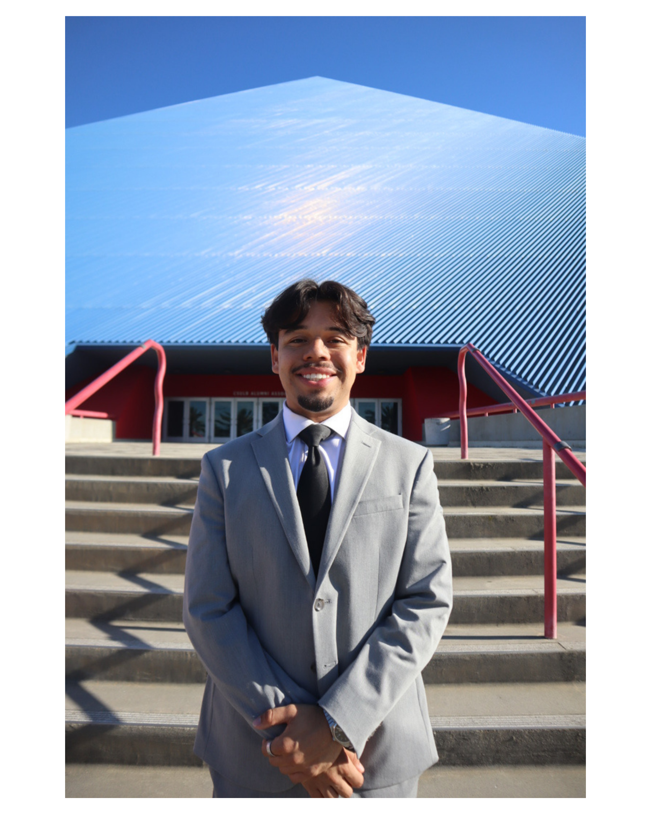 Young man smiling outdoors in a grey suit, standing in front of the CSULB Pyramid under a clear blue sky.