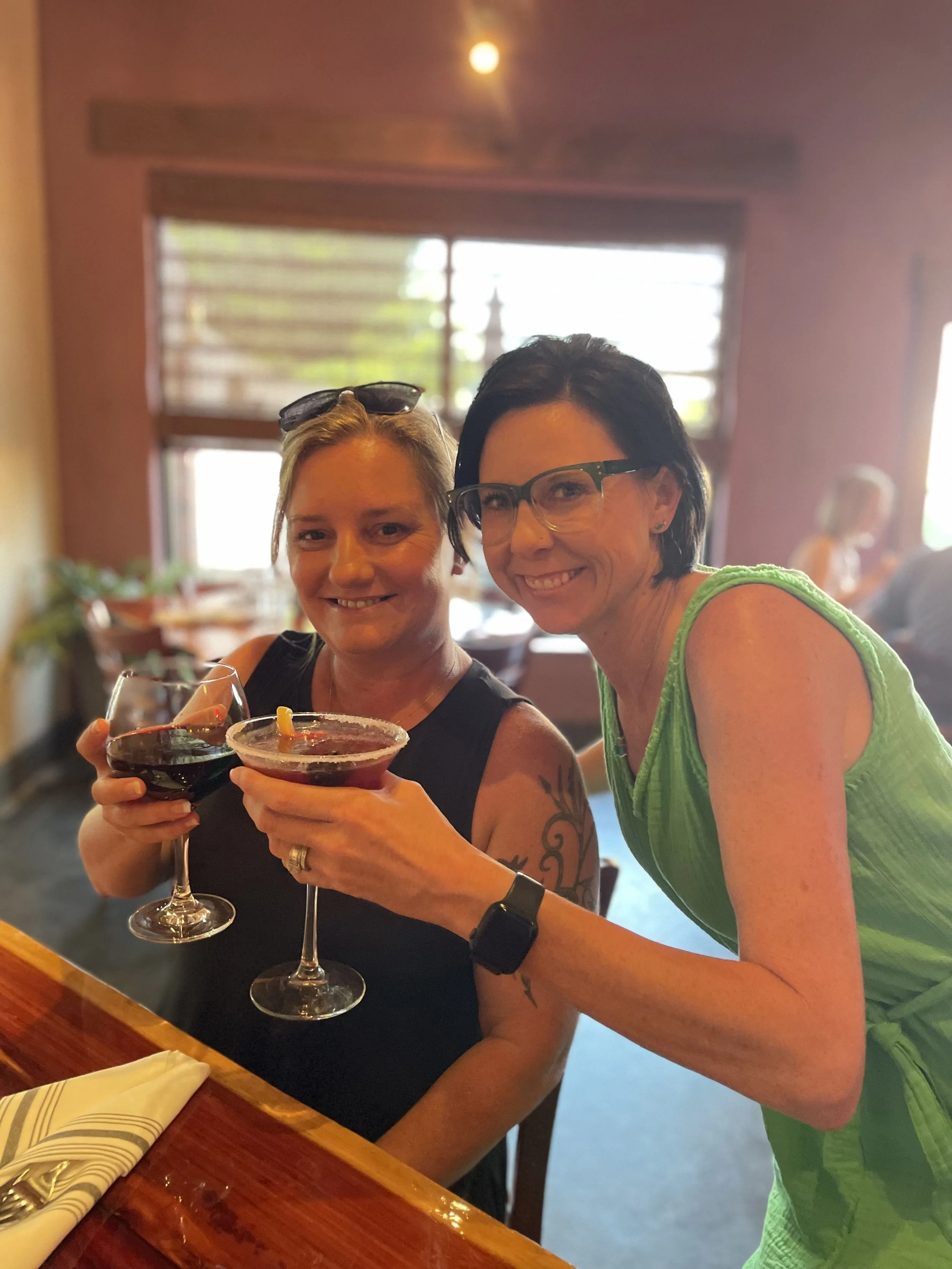 Two women smiling and holding wine glasses in a warmly lit restaurant with a large window in the background.
