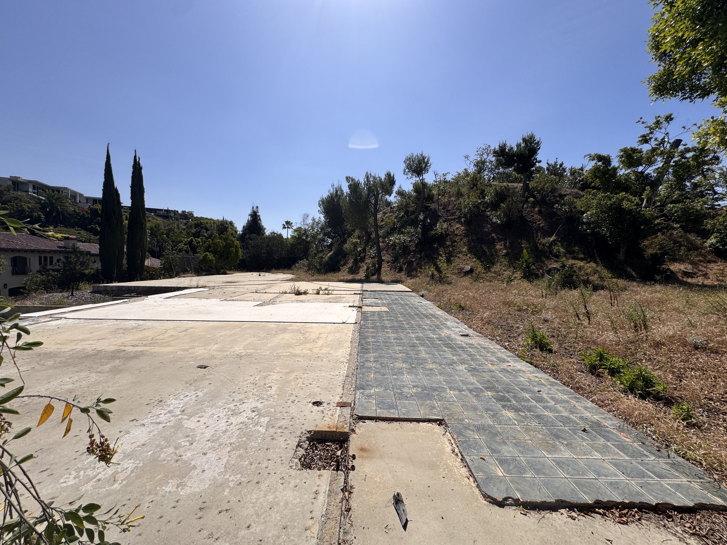 Partial sidewalk with some tiles missing, dry grass and bushes along a hill on the right side, and a blue sky with sunlight overhead.