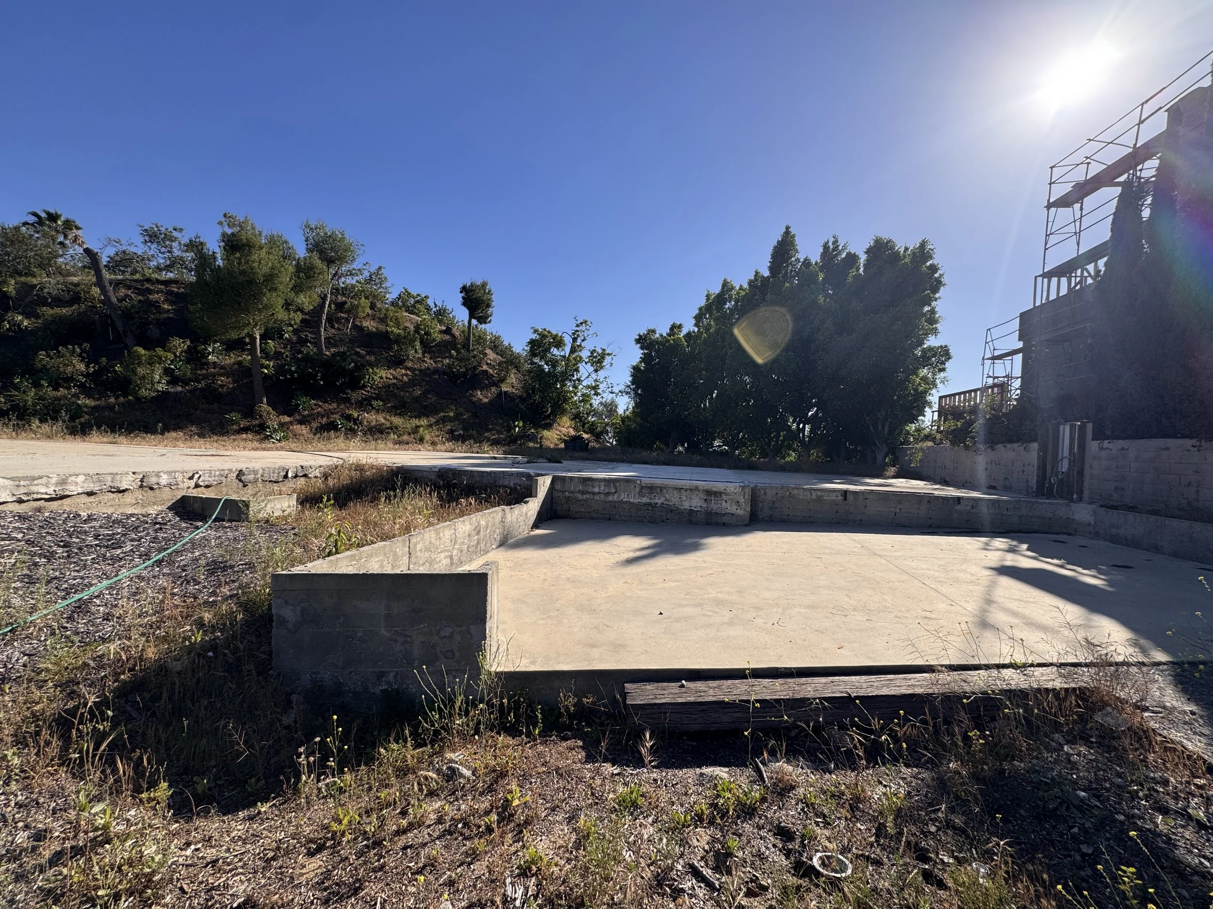 Empty concrete foundation with surrounding dirt and grass, a hillside with trees on the left, a large tree on the right, and a building with scaffolding in the upper right corner. The sky is clear with bright sunlight.