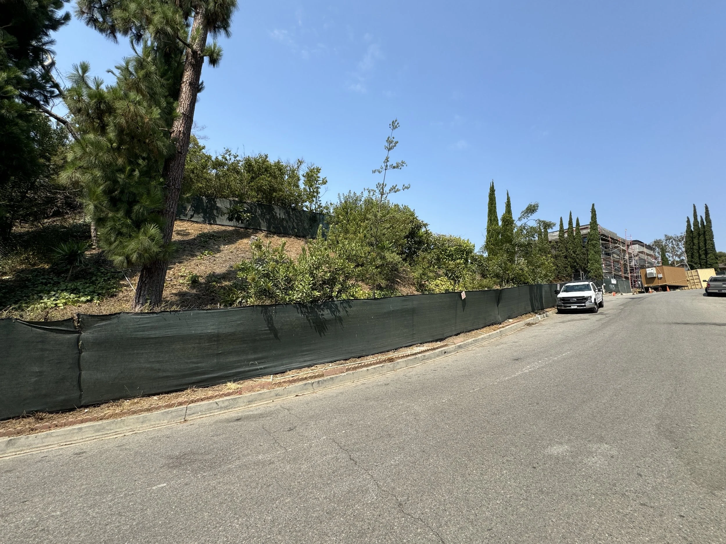 A hillside with several trees and shrubs behind a black construction fence, along a sloped street with parked cars and a building under construction in the distance.