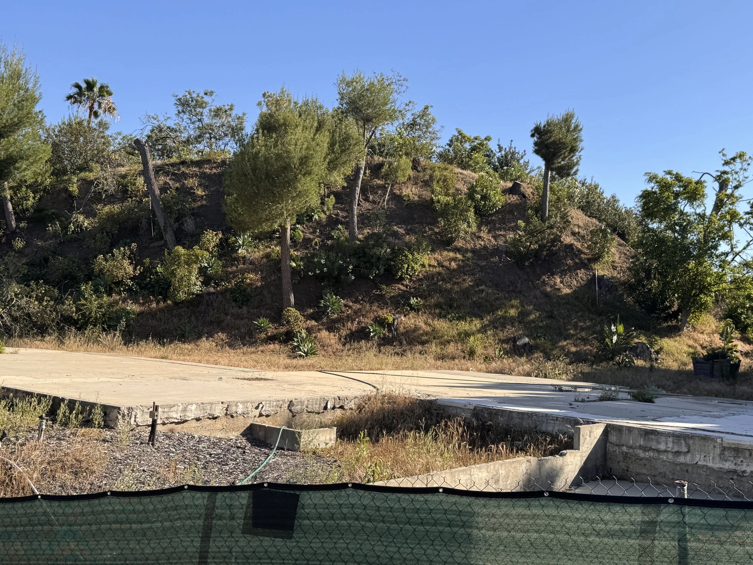 A hillside with sparse vegetation, trees, and shrubs, with a concrete slab and construction materials in the foreground under a clear blue sky.