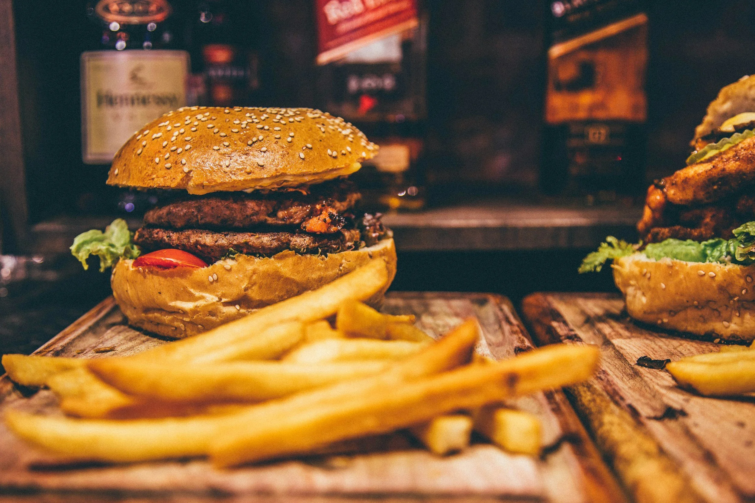Close-up of a cheeseburger with sesame seed bun, lettuce, tomato, and a beef patty, served with French fries on a wooden tray, in a dimly lit burger restaurant.
