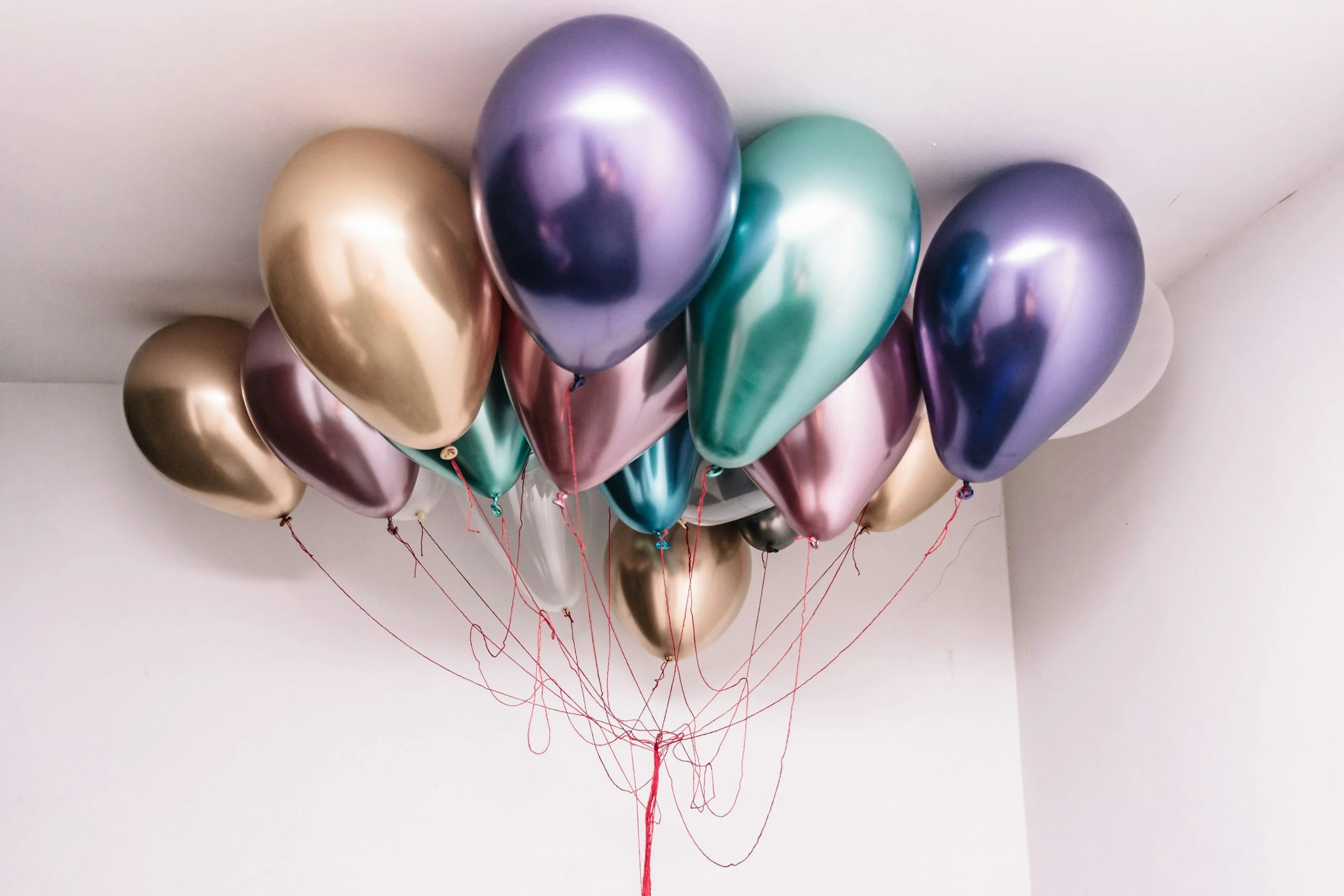 A bunch of metallic-colored balloons in gold, purple, green, and white, floating near the ceiling, with red strings hanging down.
