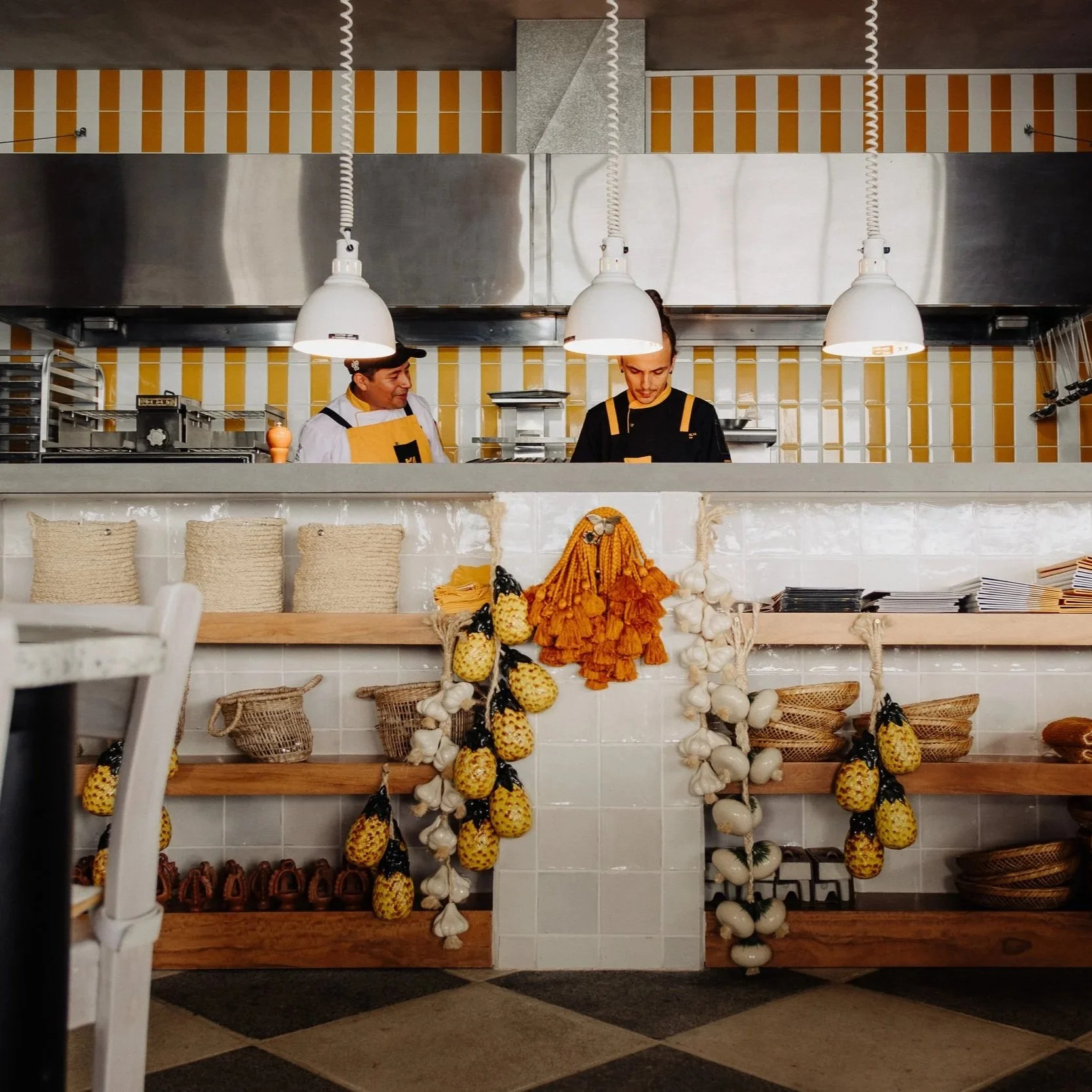 Interior view of a restaurant kitchen with two chefs working behind a counter decorated with hanging strings of pineapples and garlic. The background features yellow and white tiled walls, stainless steel appliances, and shelves with baskets and plates.