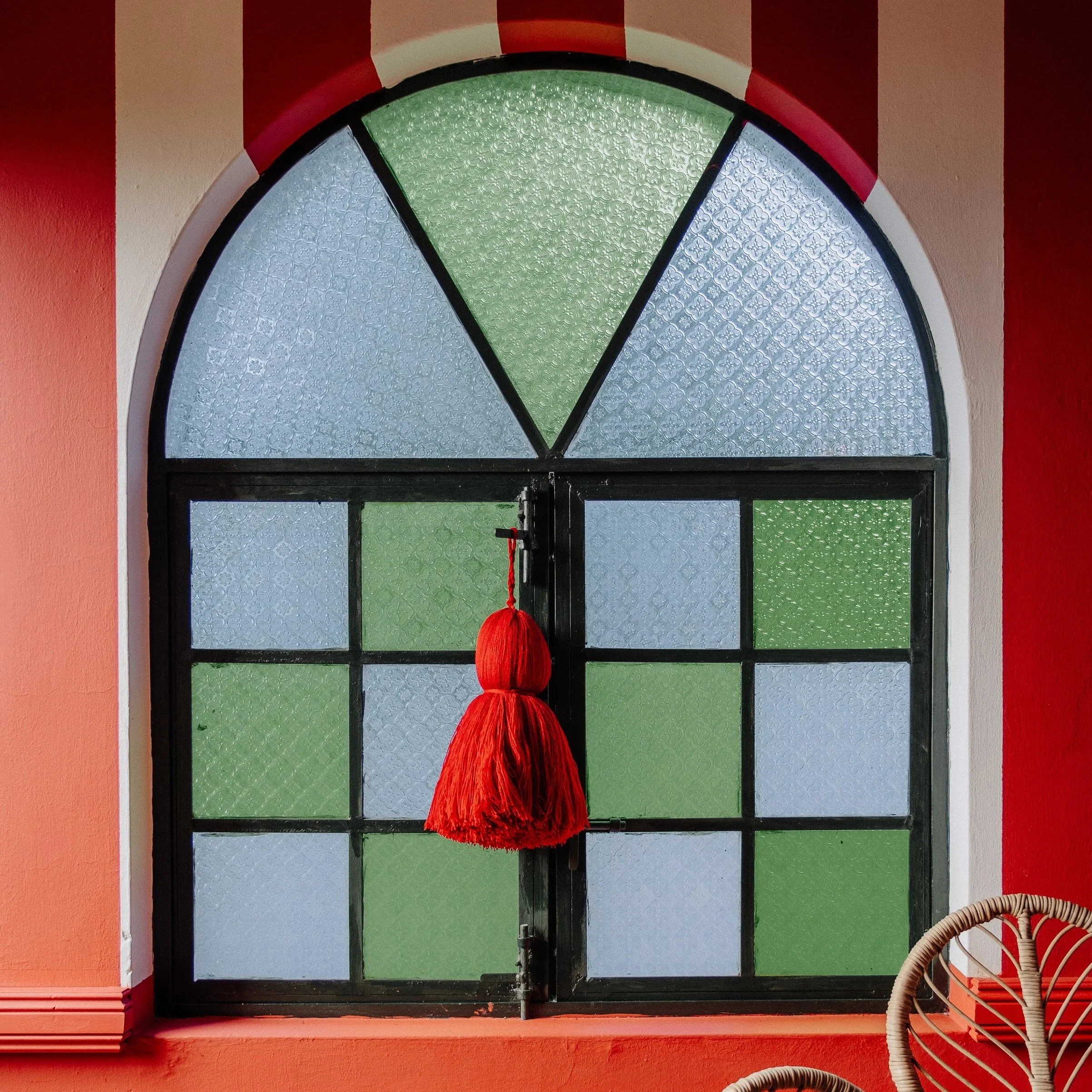 Colorful stained glass window with green, gray, and blue panels, framed by a black border, and decorated with a red hanging ornament, set in a reddish-orange wall.