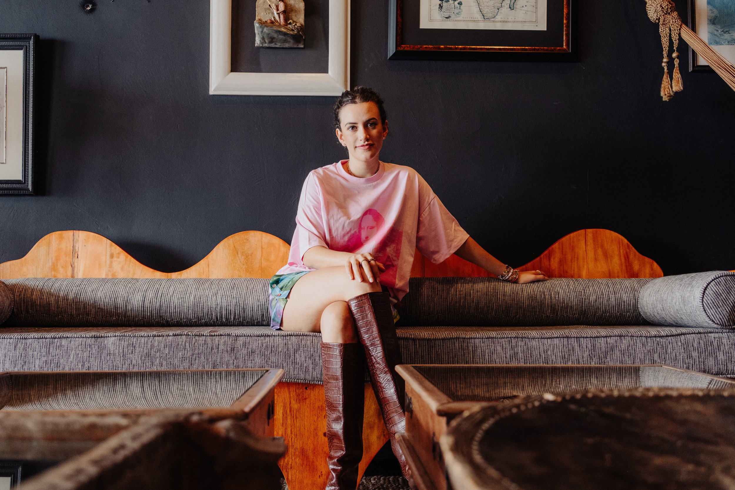 A woman with dark hair tied back, wearing a pink T-shirt with a graphic of a woman's face, colorful shorts, and tall boots, sitting on a patterned sofa in a room with framed art and a dark wall.