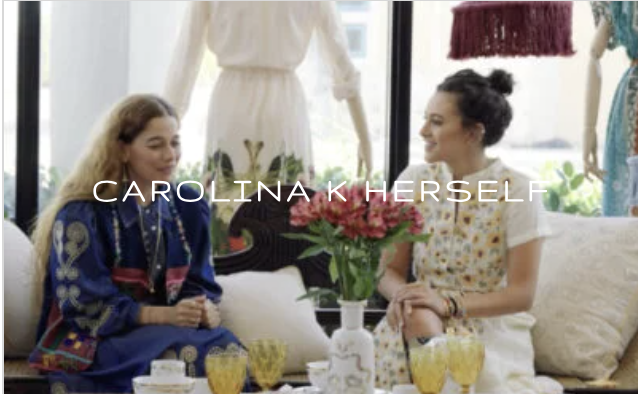 Two women sitting on a couch having a conversation in a boutique or showroom with mannequins and dresses in the background, surrounded by decorative items and tea cups.
