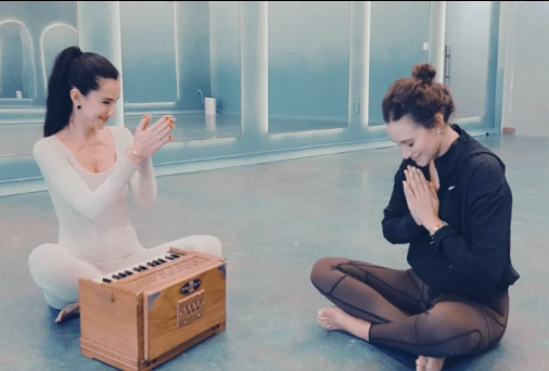 Two women sit cross-legged on the floor, one playing a small wooden harmonium and the other with hands in a prayer position, both smiling in a bright, modern room.