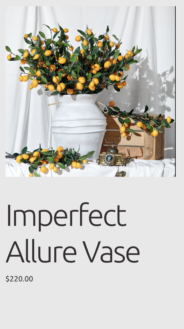 A white, imperfect Allure Vase filled with yellow-orange citrus fruits and green leaves, set on a white table with a wooden box and vintage camera nearby, against a white backdrop.